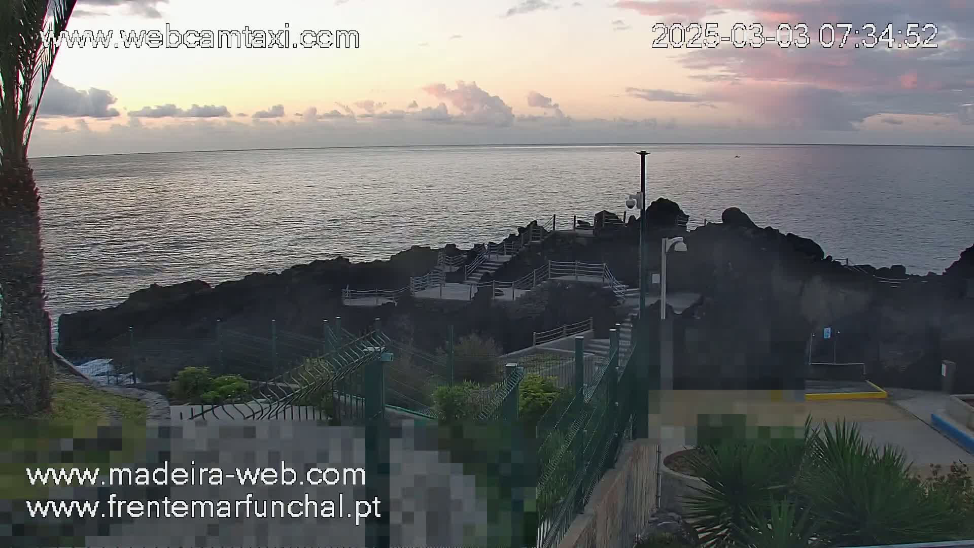 A rocky coastal area with stairs leading down to the ocean, under a partly cloudy, sunrise sky.