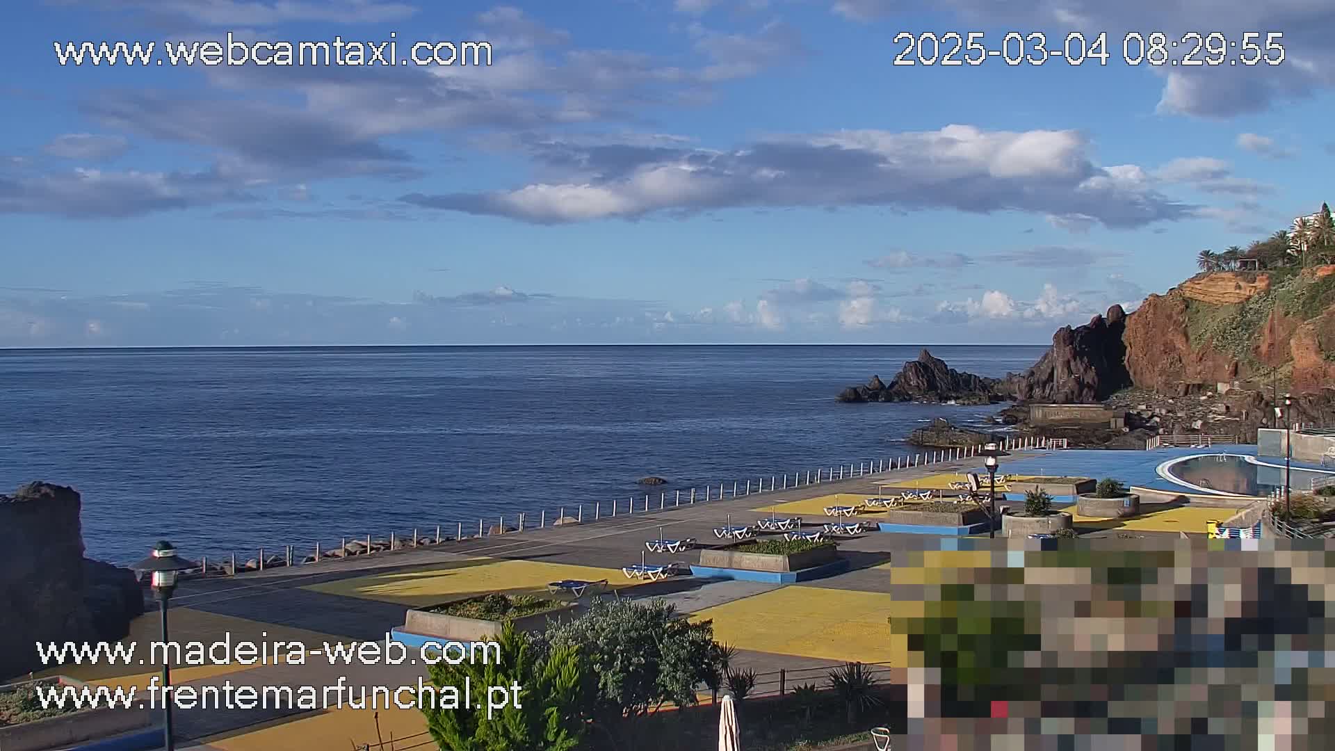 A seaside paved area with lounge chairs and a swimming pool overlooks a calm ocean under a partly cloudy sky.