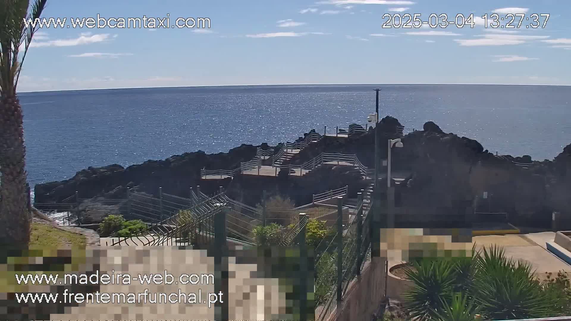 A rocky coastline with a stairway leading down to the ocean is visible under a mostly clear, sunny sky.