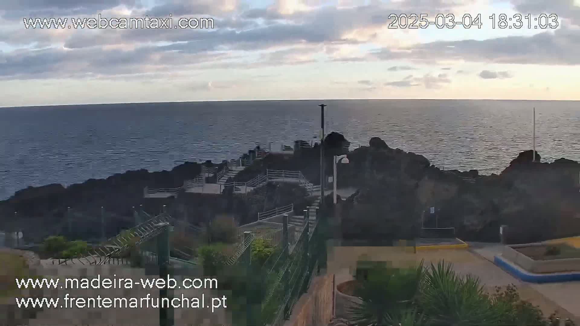 A rocky coastal area with stairs leading down to the ocean under a partly cloudy sky.
