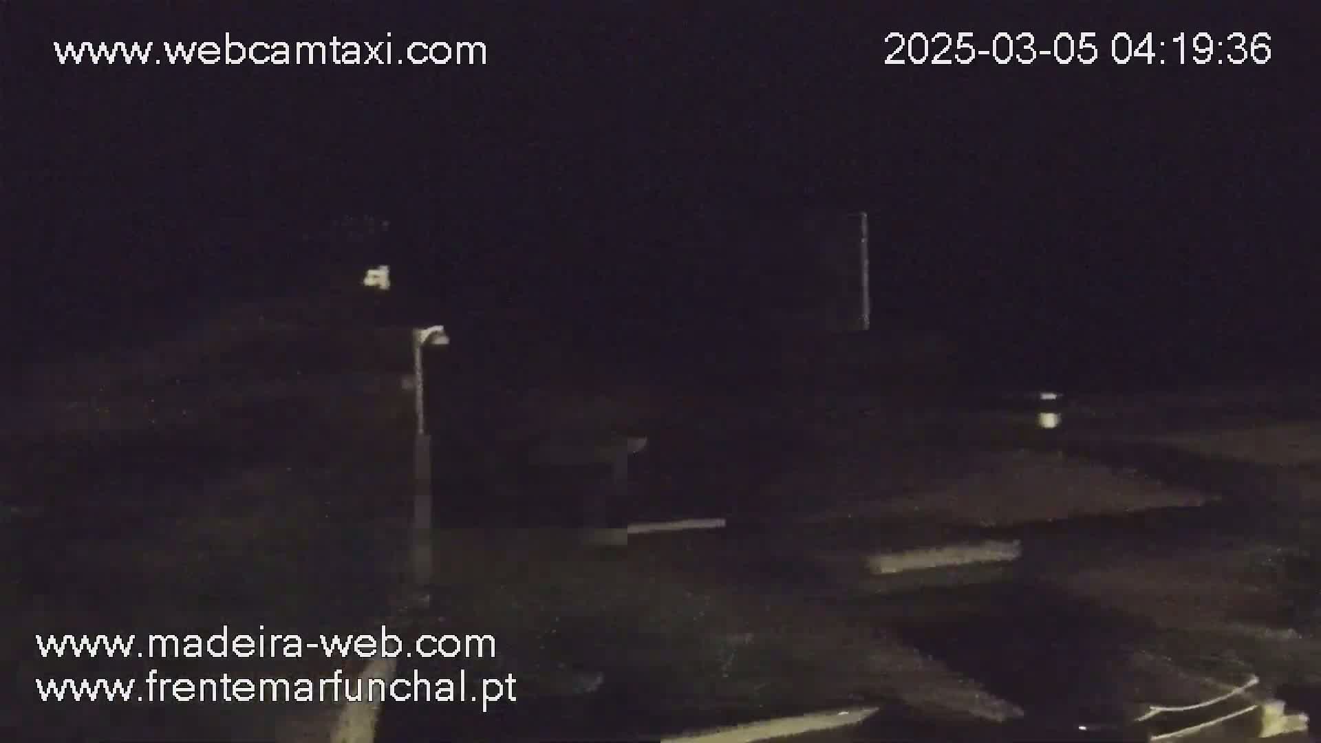 A nighttime, dark outdoor scene shows a sparsely lit area with some structures and a lamppost, possibly a parking lot or roadway.