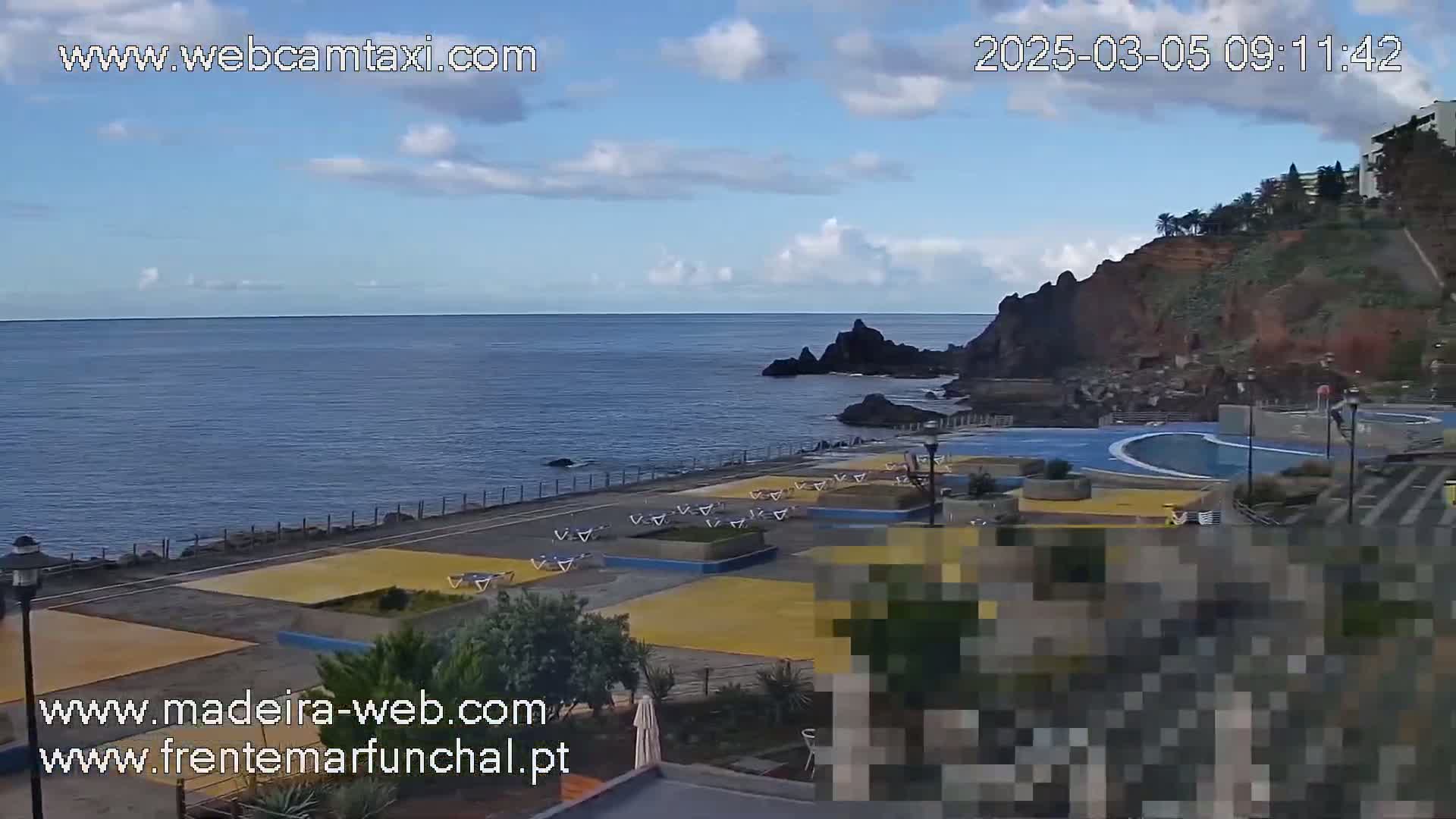 A partly sunny day overlooking a coastal area with a paved walkway, an empty swimming pool, and lounge chairs situated near dark cliffs and a calm ocean.