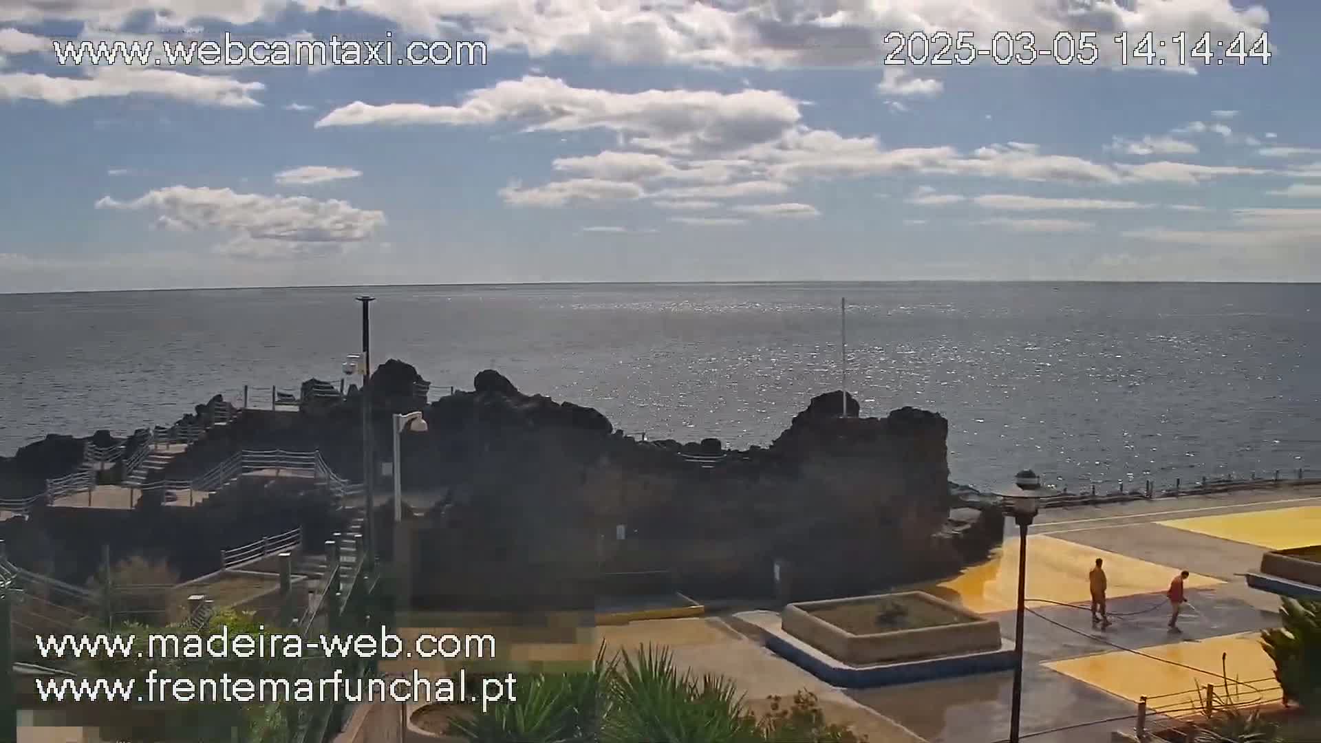 A partly cloudy day shows a rocky coastline with a calm ocean in the background, and two people are painting yellow stripes on a paved area next to the water.
