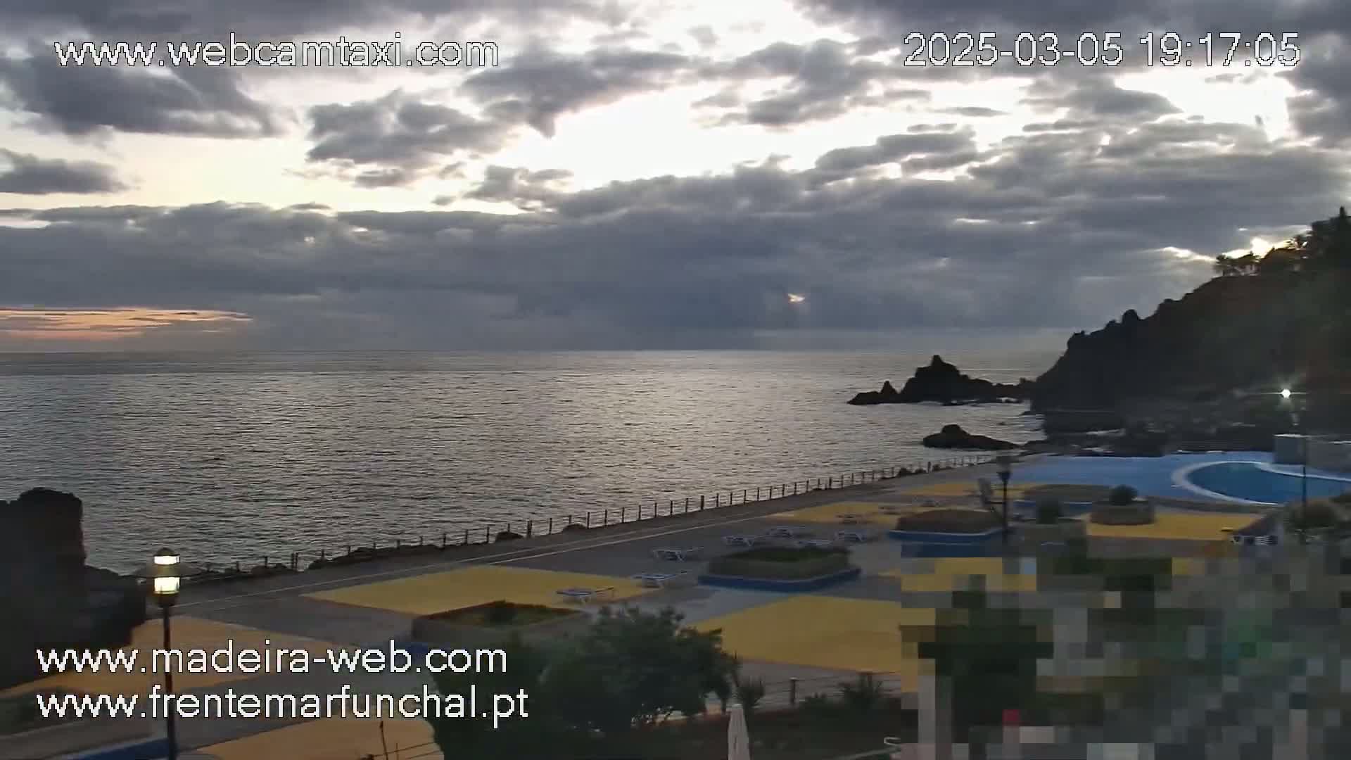 An overcast sky over a calm ocean meets a rocky coastline with a paved area containing lounge chairs and a swimming pool.
