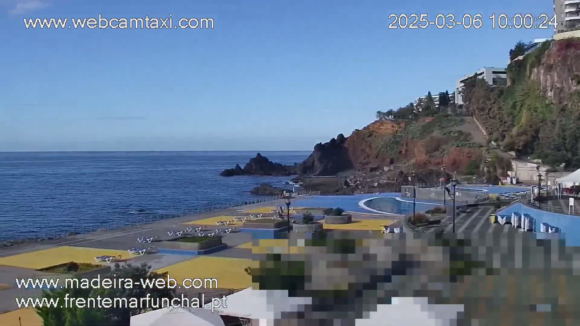 An outdoor scene shows a calm ocean, a rocky coastline with buildings on a cliff, and a public pool area with yellow and blue tiled surfaces.