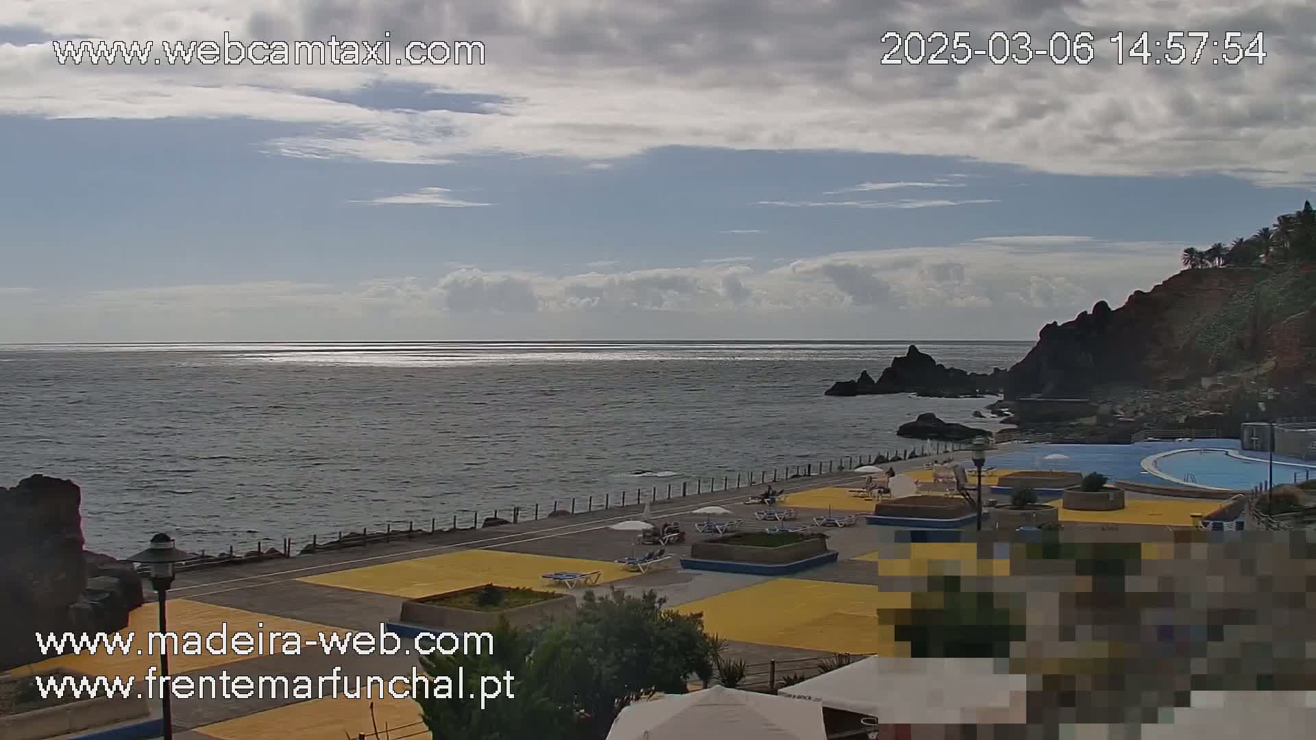 An outdoor scene shows a partly cloudy sky over a dark-blue ocean with a rocky shoreline featuring a pool area with lounge chairs and umbrellas.