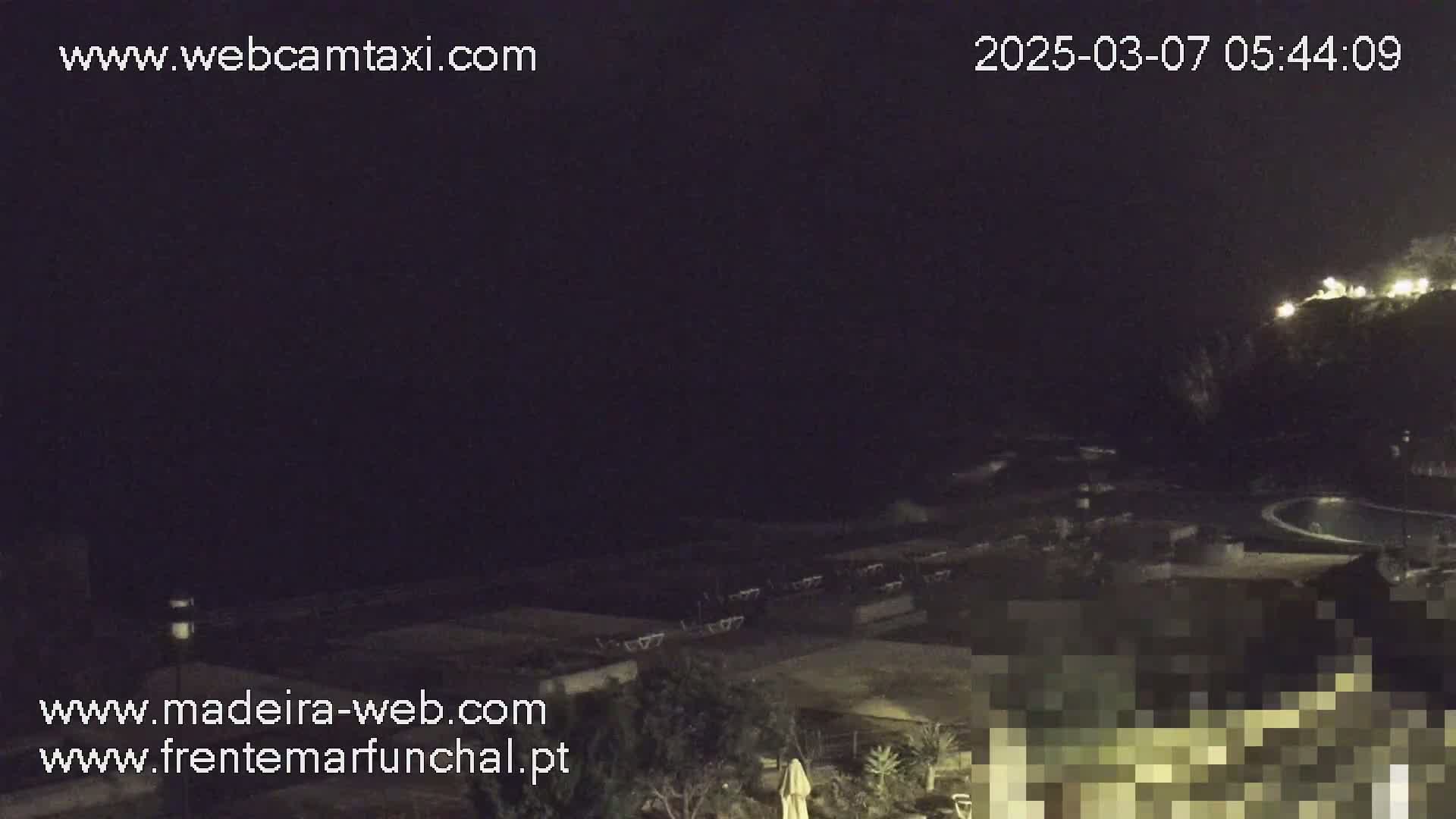 A nighttime, outdoor view of a dark ocean and coastline, featuring a pool and some darkened outdoor furniture.