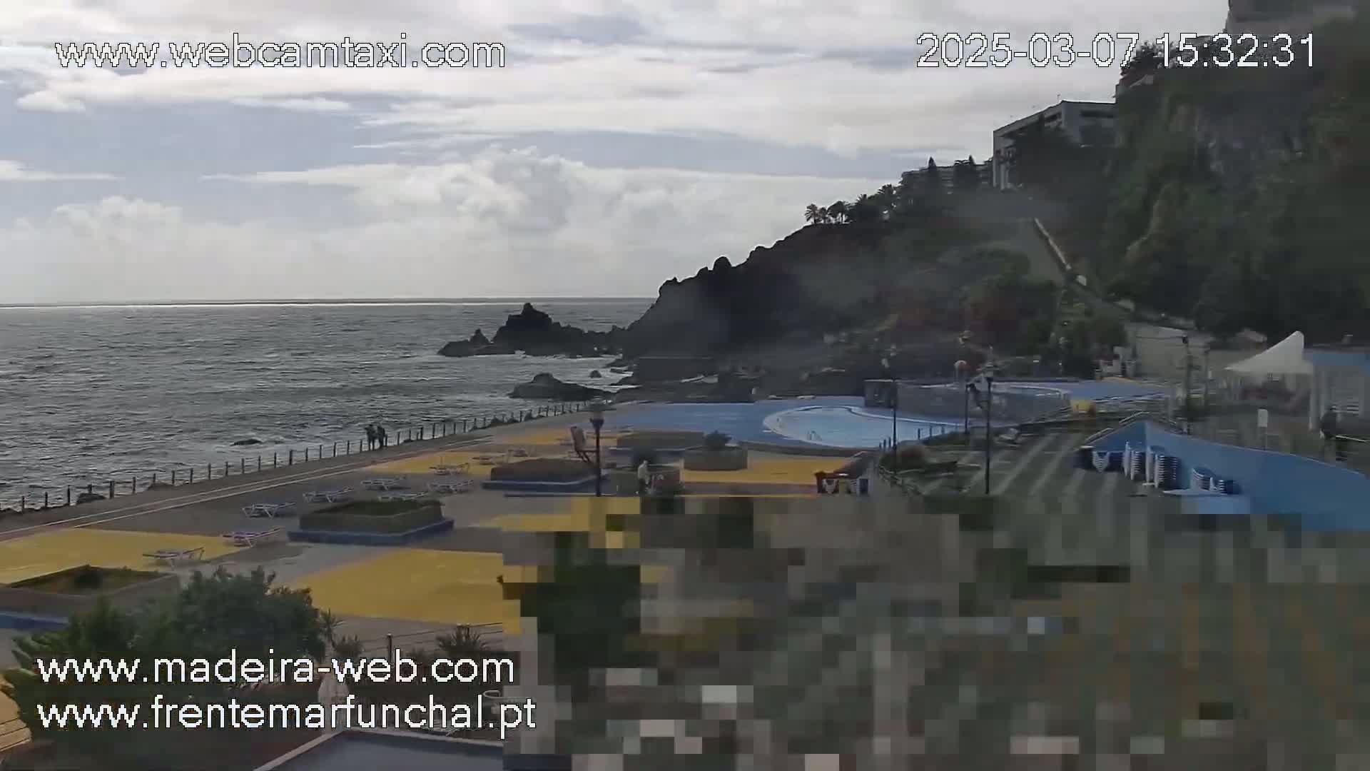 A coastal outdoor scene shows a calm ocean, rocky cliffs with buildings on top, and a paved area with pools and lounge chairs under a mostly cloudy sky.