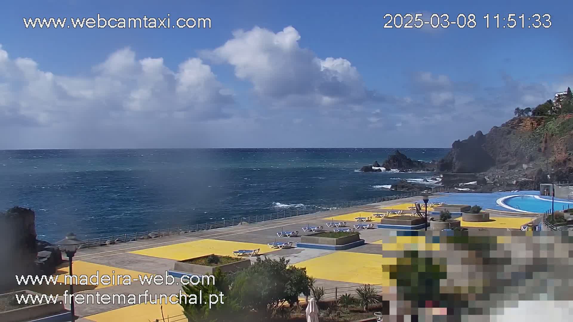 A partly cloudy day shows an outdoor pool area next to a rocky coastline and a blue ocean with small waves.