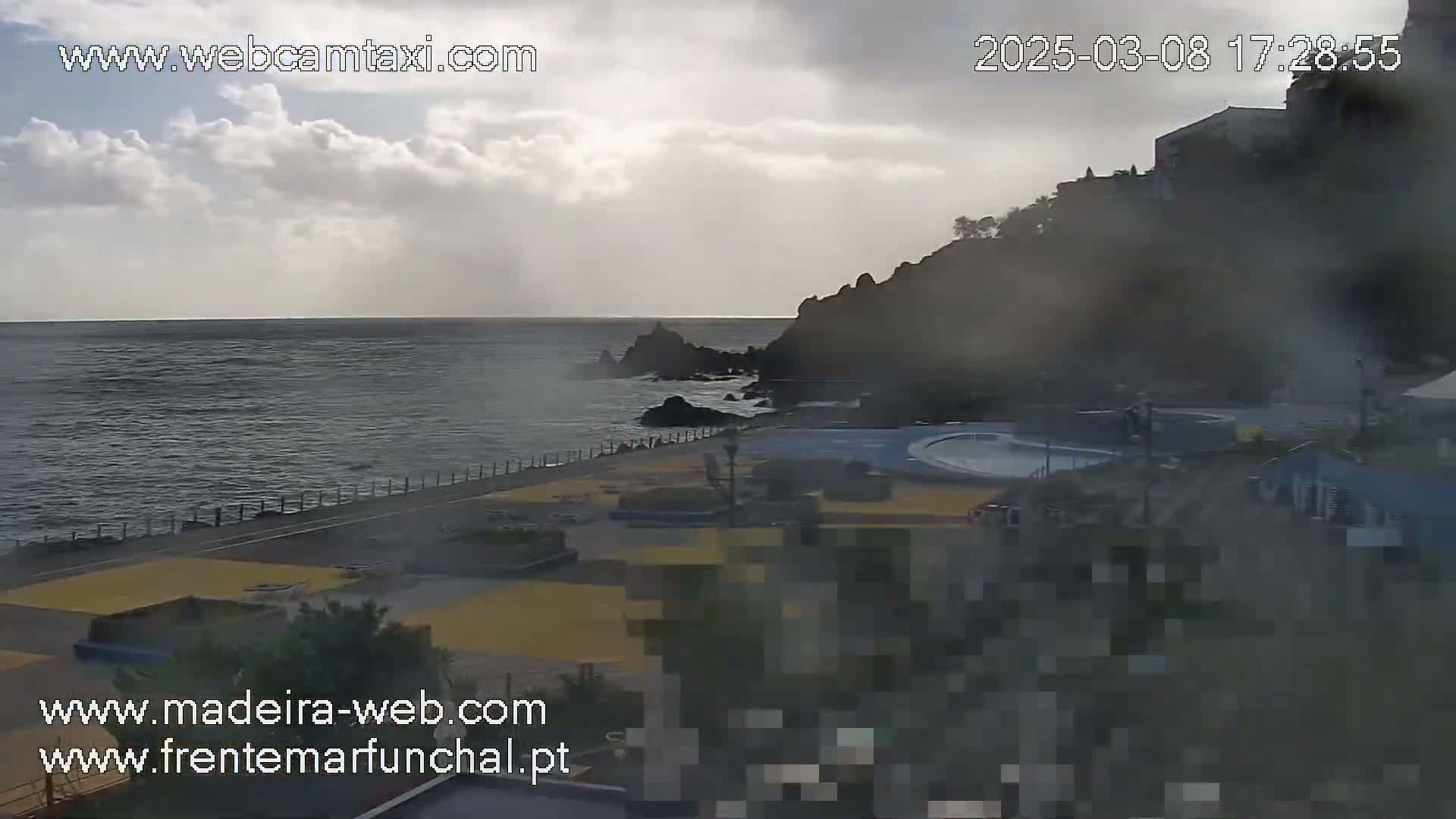 A mostly cloudy day overlooks a coastal area with a paved walkway, a dark-colored rocky cliff, and a body of water.