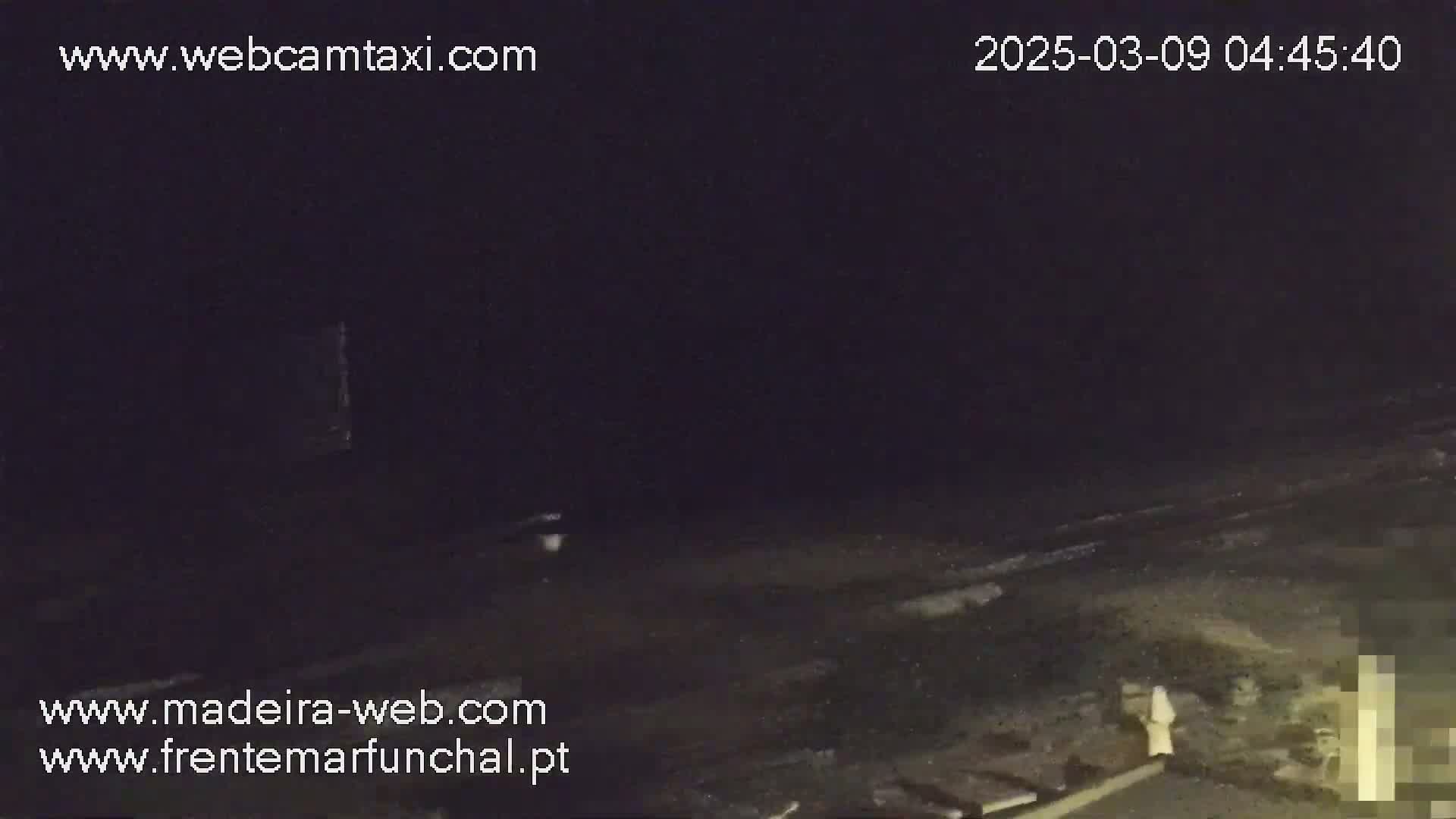 A nighttime, dark outdoor scene shows a mostly empty, dark beach with a single, light-colored figure standing near some structures in the lower right corner.