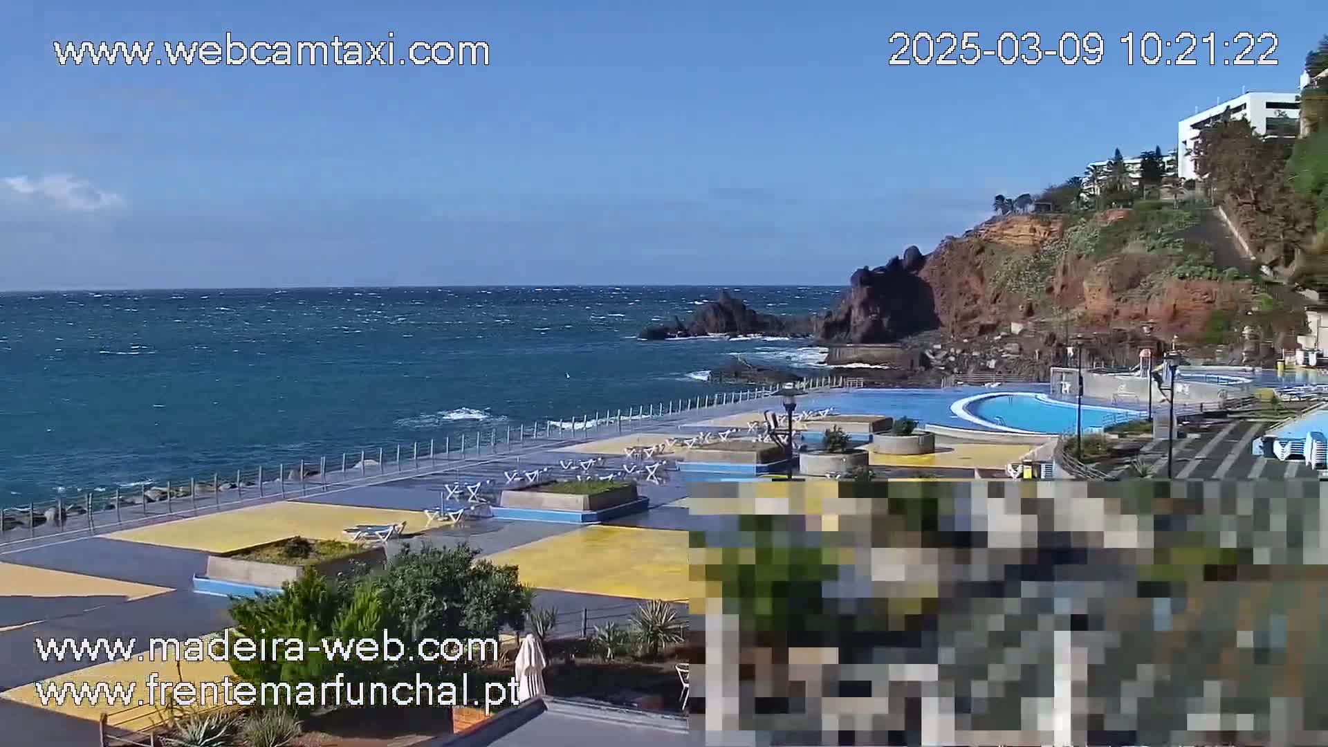 A windy seaside outdoor scene shows a paved area with lounge chairs and an outdoor pool complex next to a rocky coastline and ocean.