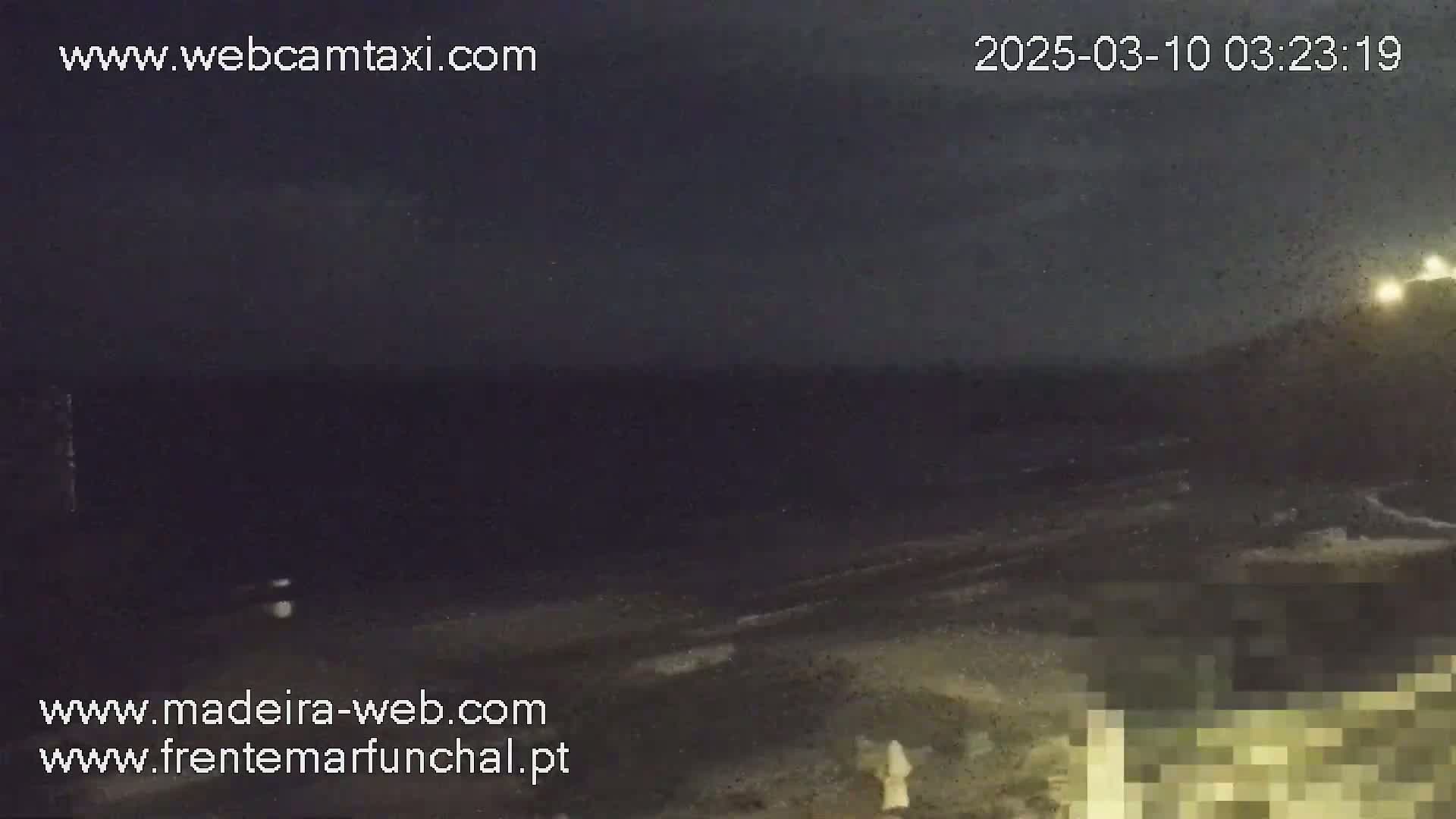 A dark, nighttime coastal scene shows a dark ocean with small waves lapping a dark beach under a cloudy sky.