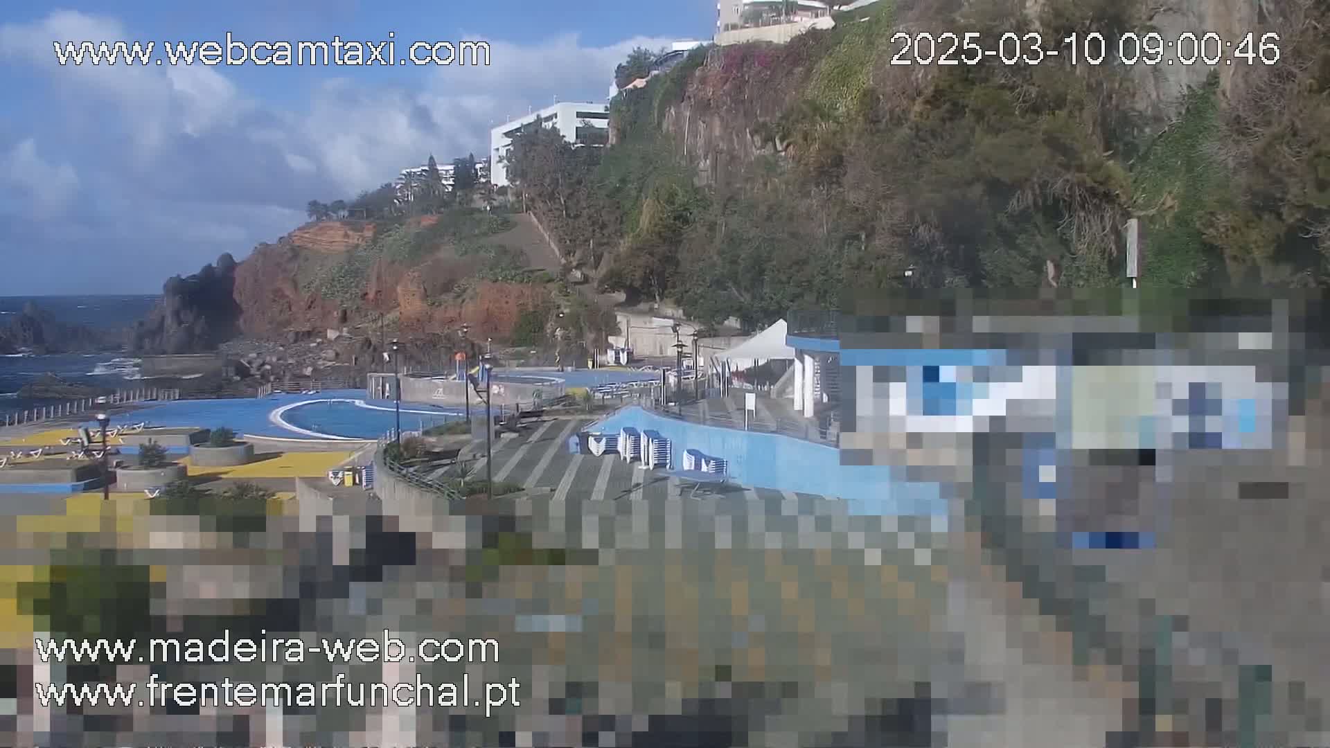 An outdoor oceanside scene on a partly cloudy day shows a pool area with buildings and cliffs in the background.