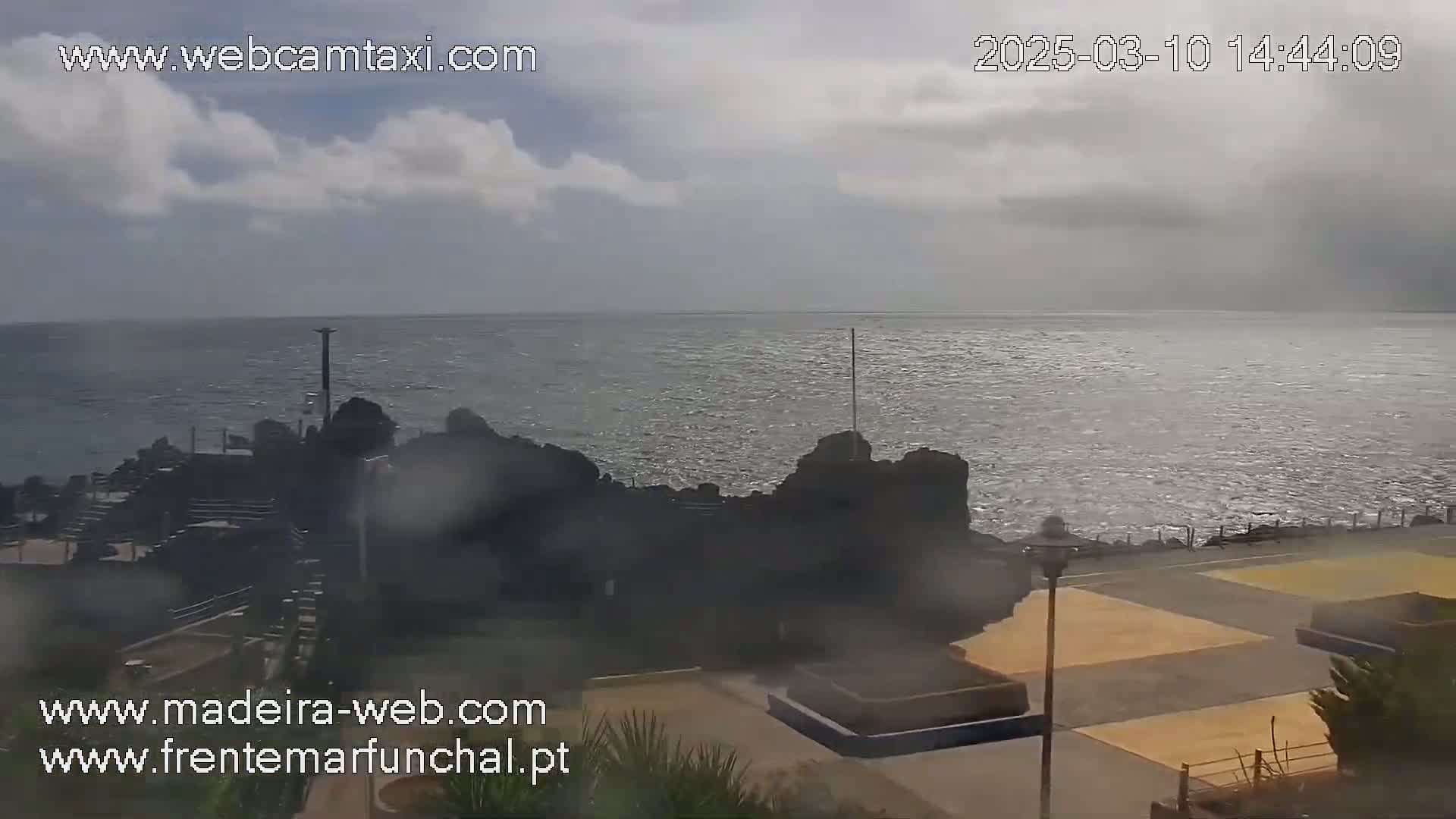 A partly cloudy day shows a rocky coastline meeting a somewhat choppy ocean, with a paved area and some landscaping visible in the foreground.