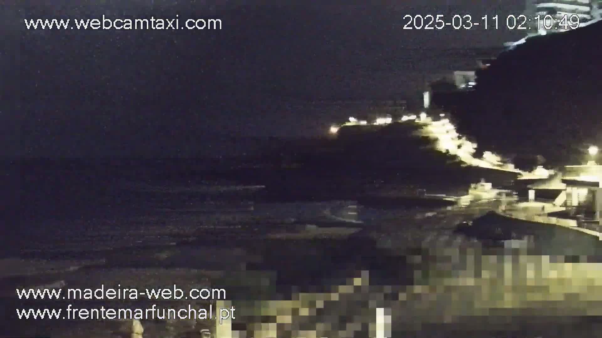 A nighttime coastal scene shows dark water, a dark cliff face with lights on top, and a dark shoreline.