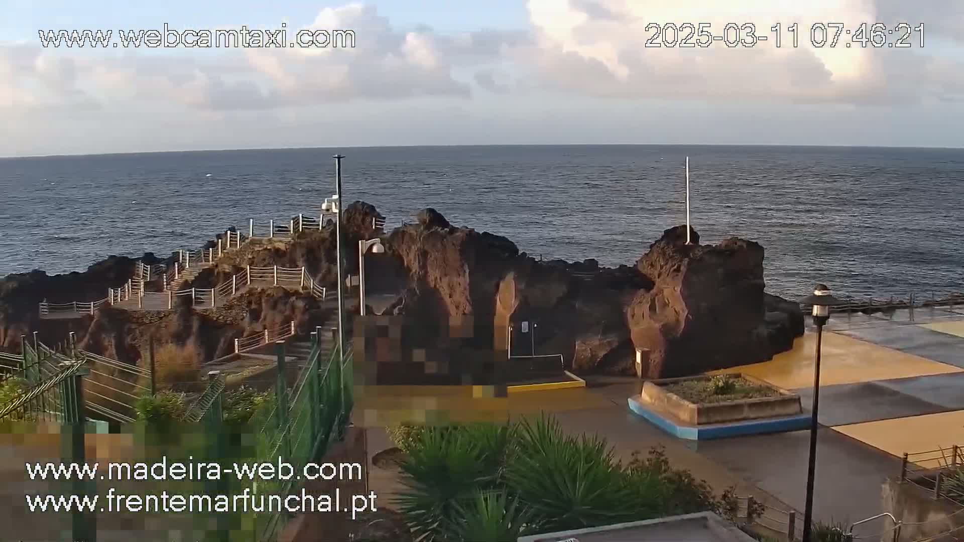 A rocky coastal area with a paved walkway and a calm ocean under a partly cloudy sky.