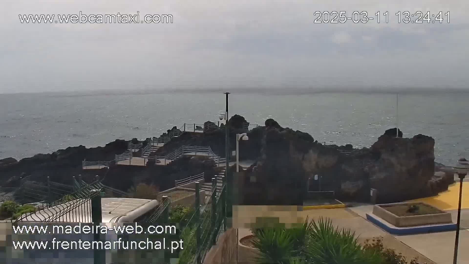 A rocky outcrop juts into a gray, overcast ocean, with paved pathways and a small plaza visible nearby.