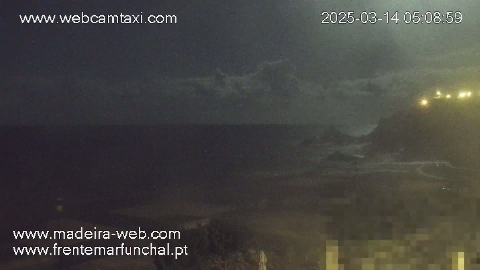 A nighttime, overcast view of a dark ocean meeting a landmass with lights visible on a hill in the distance.