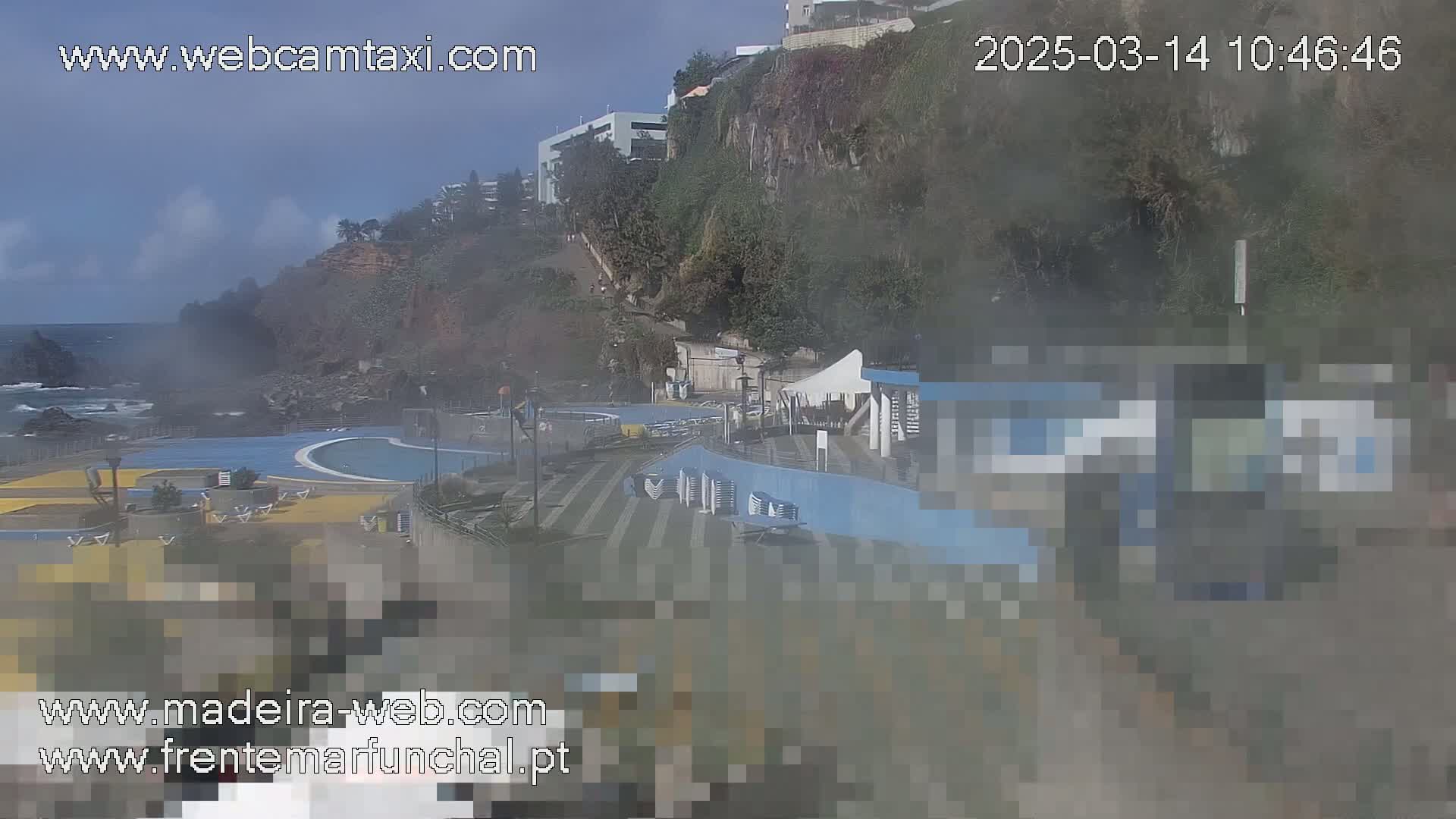 An outdoor view on a slightly hazy day shows a seaside pool complex nestled against a cliffside, with the ocean visible in the background.