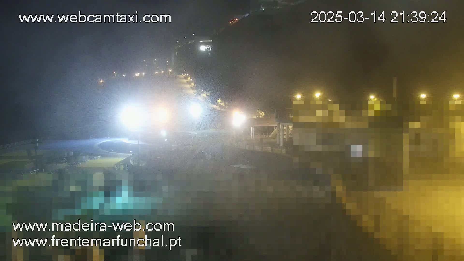 A nighttime view of a coastal area with light rain or mist, showing illuminated buildings and a dark, wet roadway.