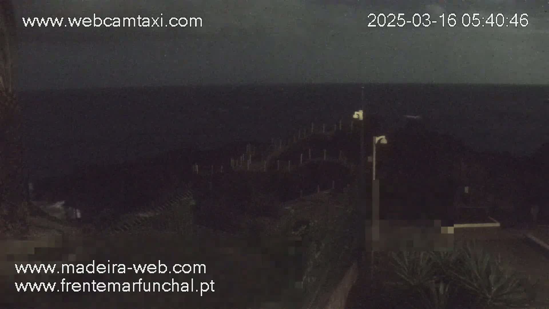 A dark nighttime scene shows a mostly obscured body of water, a shoreline with low vegetation, and some dimly lit structures.