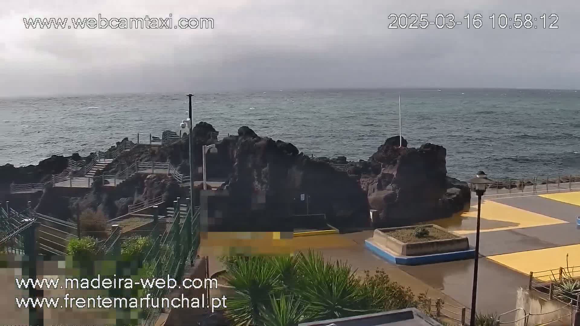 A rocky coastline with a paved area and some vegetation is visible under an overcast sky, with a somewhat choppy ocean in the background.