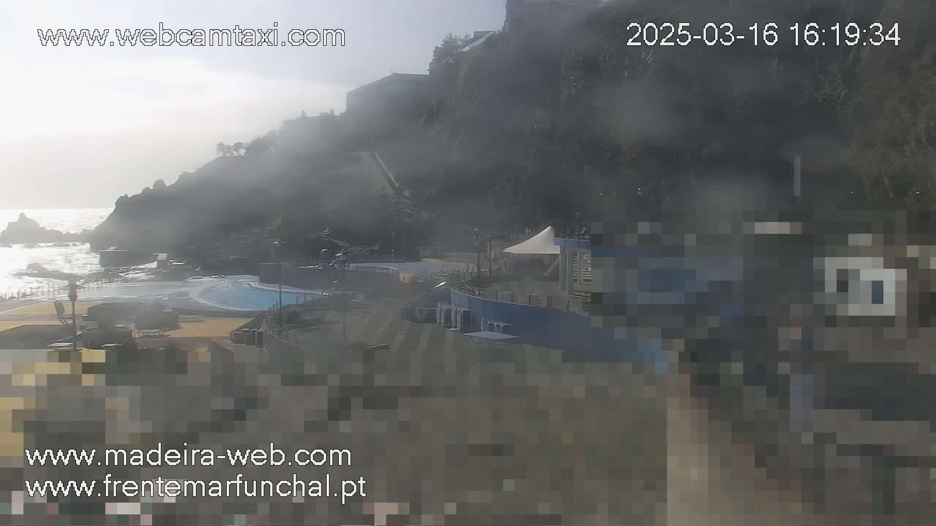 A hazy view shows a coastal outdoor swimming pool area next to a rocky cliff face, with somewhat rough ocean waves visible.