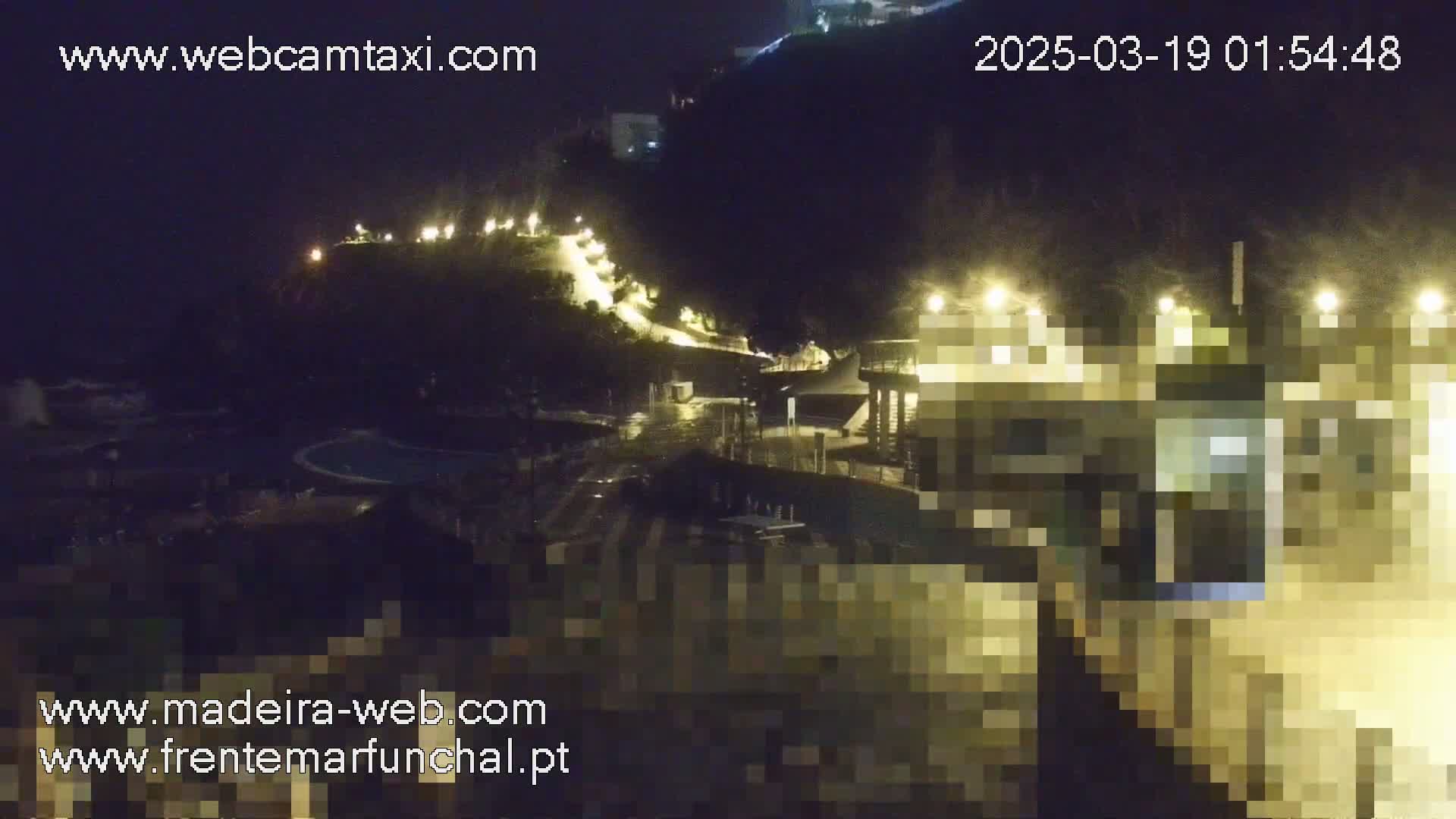 A nighttime view of a coastal area with illuminated buildings and a path winding up a hill, under cloudy conditions.