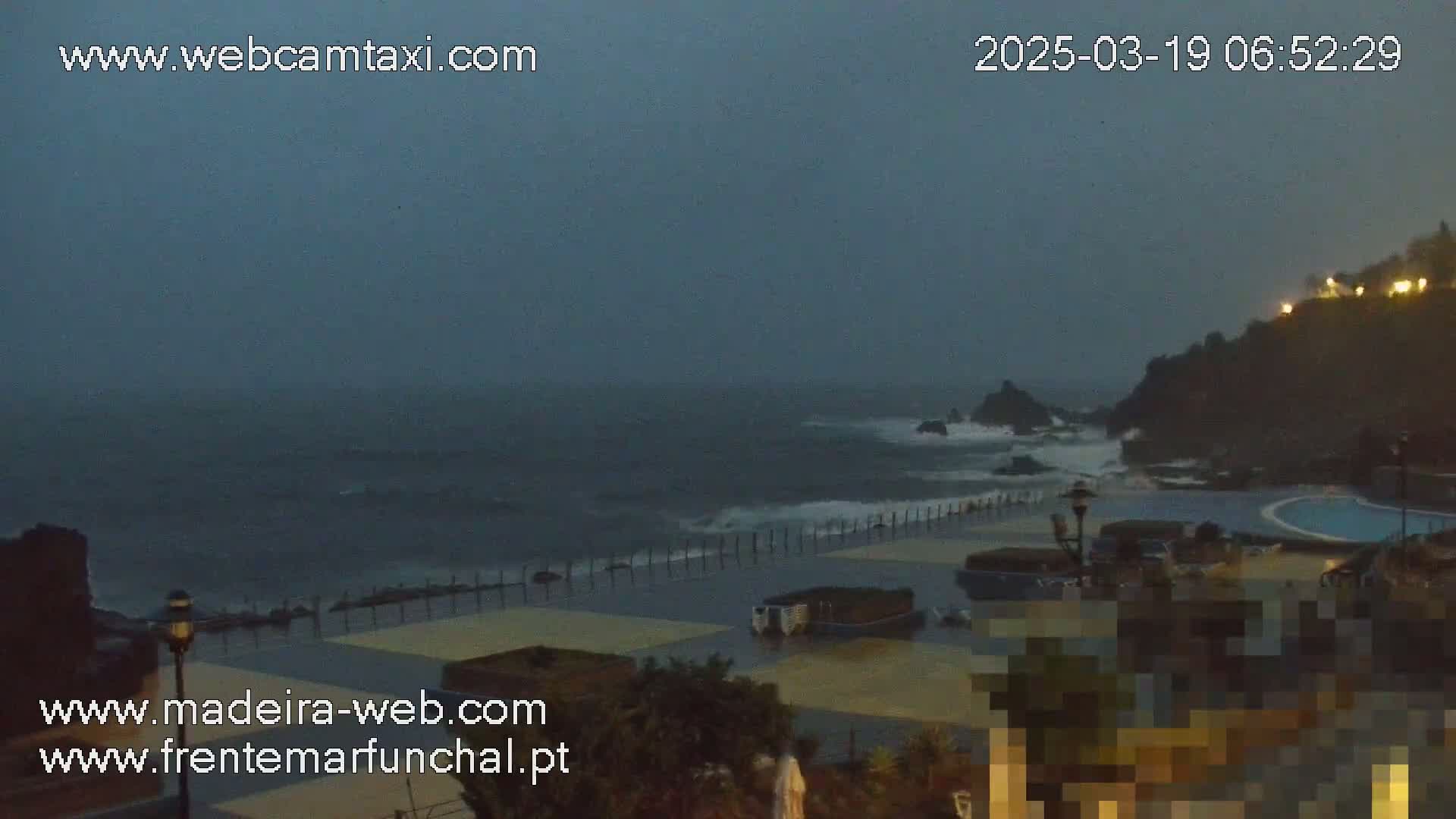 A stormy, dark gray ocean crashes against a seawall next to a mostly empty plaza with a pool, under an overcast sky.