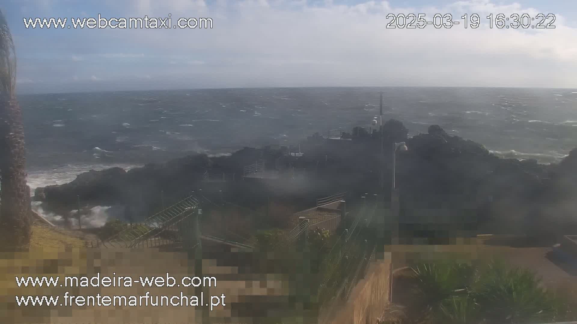 A rocky coastline is battered by a windy, choppy sea under a mostly cloudy sky.