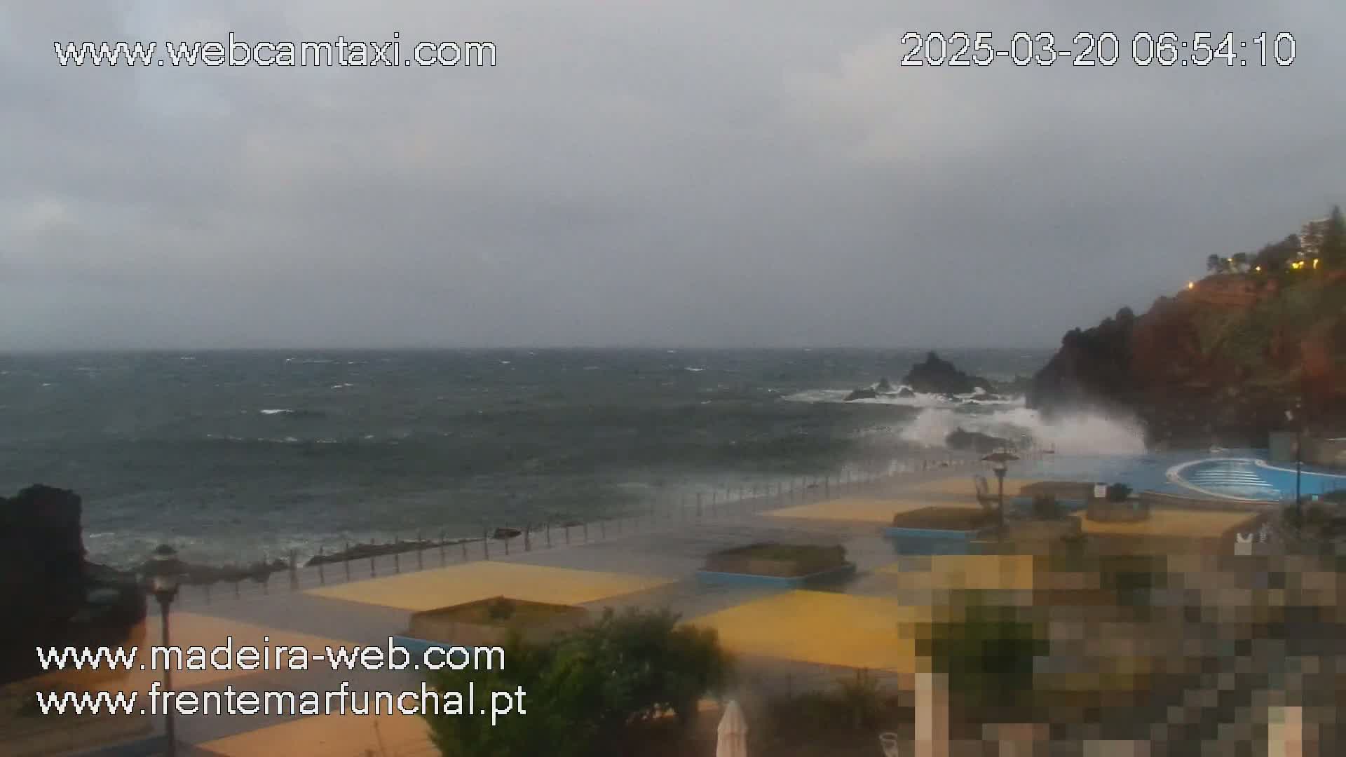 A rocky coastline with a partially flooded outdoor pool area is battered by large waves under an overcast sky.