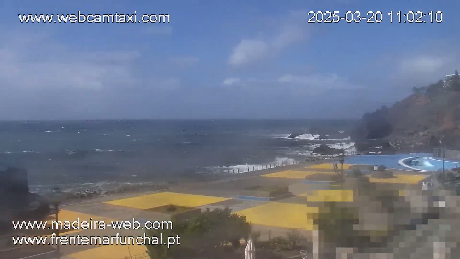 A partly cloudy day shows a rocky coastline with a wavy ocean and a pool area.