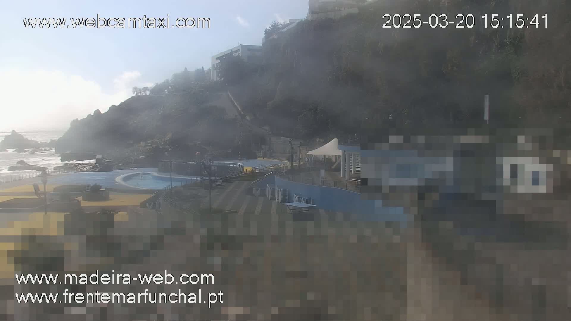 A hazy view shows an outdoor pool complex near a rocky ocean coast, with somewhat choppy waves visible.