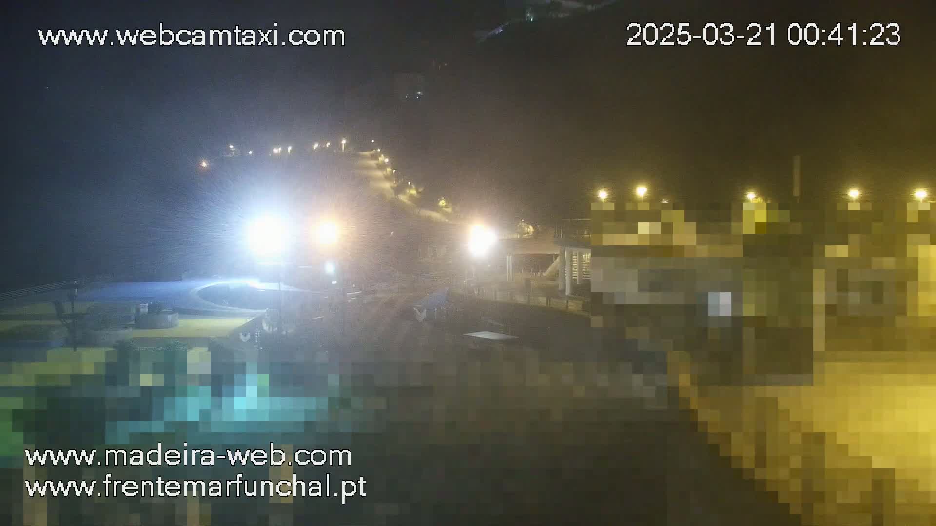A nighttime view of a rain-swept area with illuminated buildings and a pool, showing a hill with lights leading up it.
