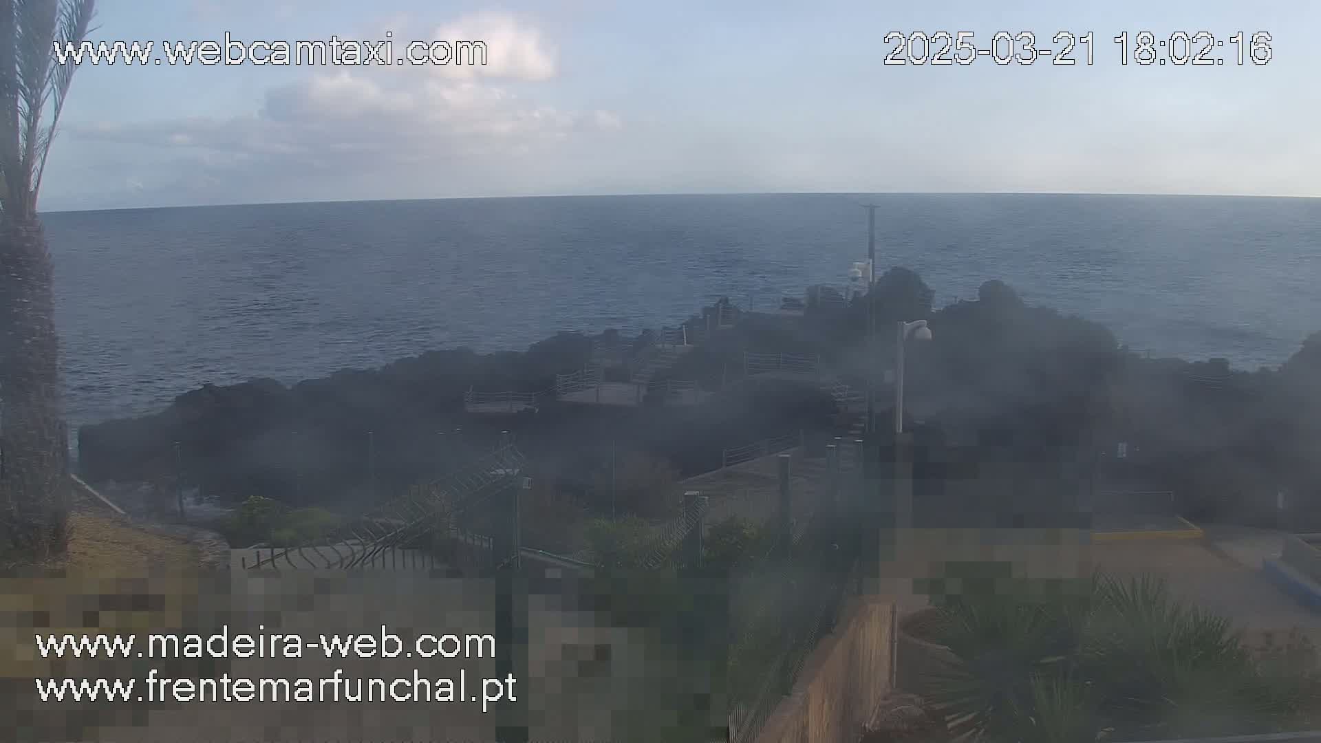 A hazy view shows a calm ocean meeting dark, rocky shoreline with some structures and vegetation in the foreground under a mostly clear sky.