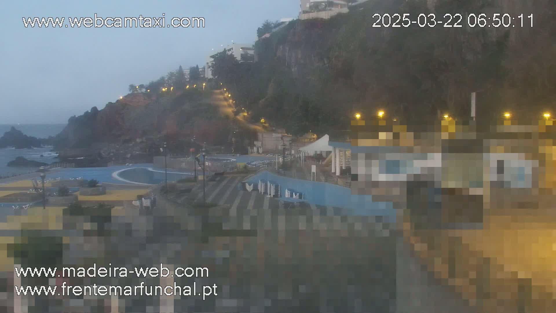 A hazy, dimly lit seaside area with a pool, buildings, and a cliffside road is seen at night.