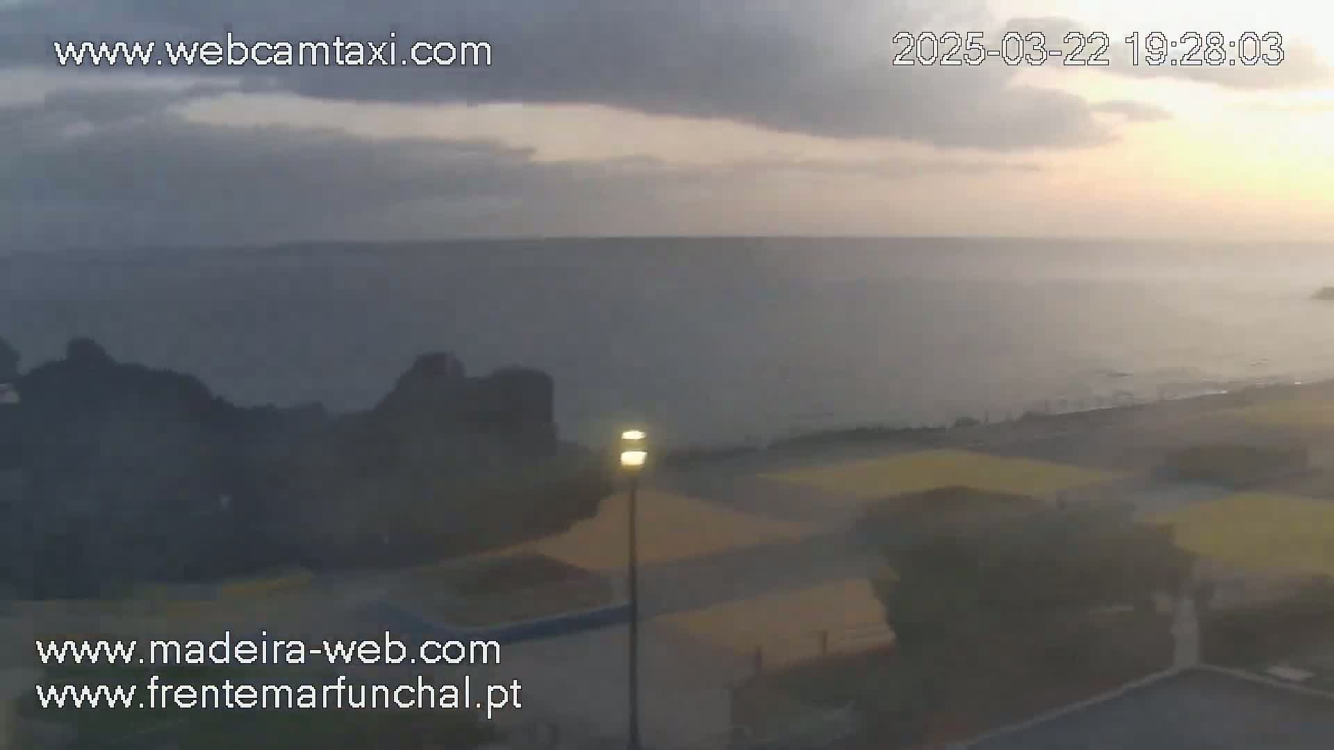 A hazy sunset over a calm ocean is viewed from a coastal area with dark rocks and a paved surface near buildings.