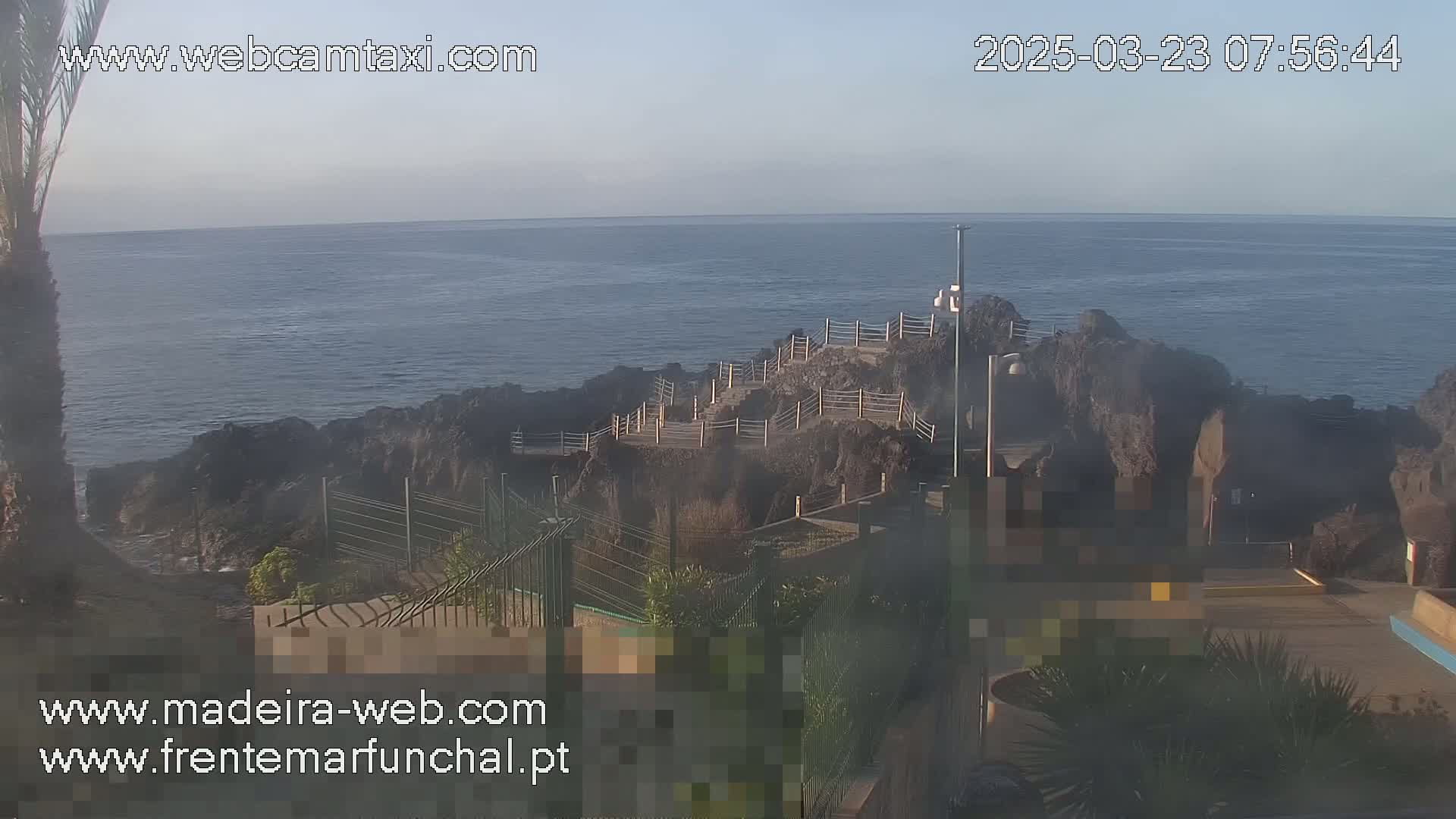 A rocky coastal area with a walkway and railing overlooking a calm ocean under a clear, light sky.