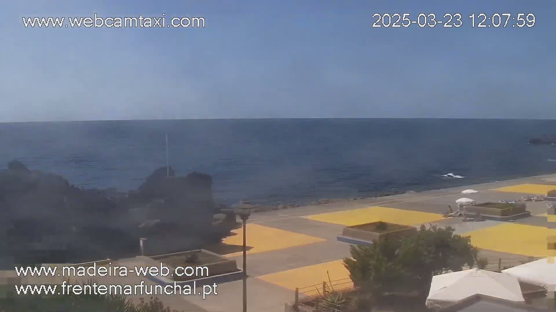 A mostly sunny day reveals a calm ocean meeting a paved beach area with a few beach umbrellas and dark volcanic rock formations.