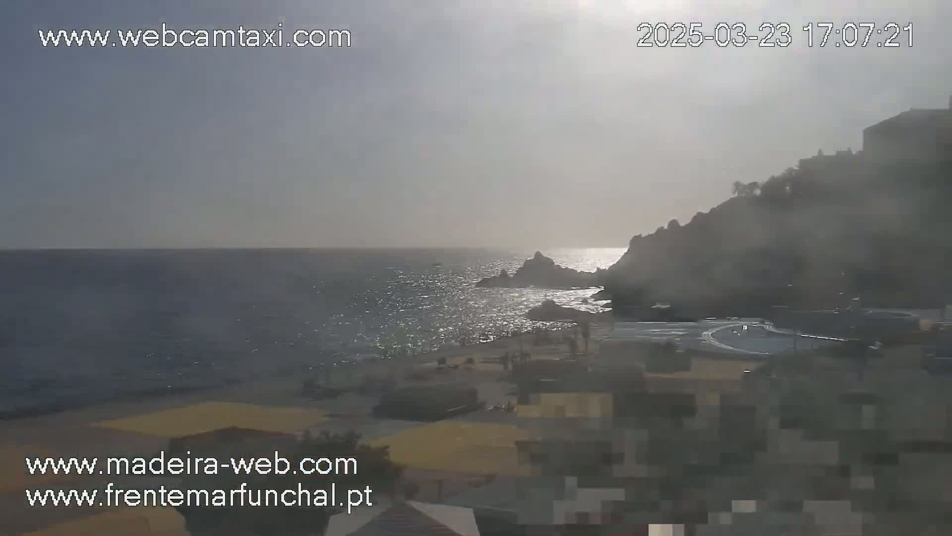 A hazy, sunny day overlooks a calm ocean meeting a rocky shore with buildings and a small beach visible in the foreground.