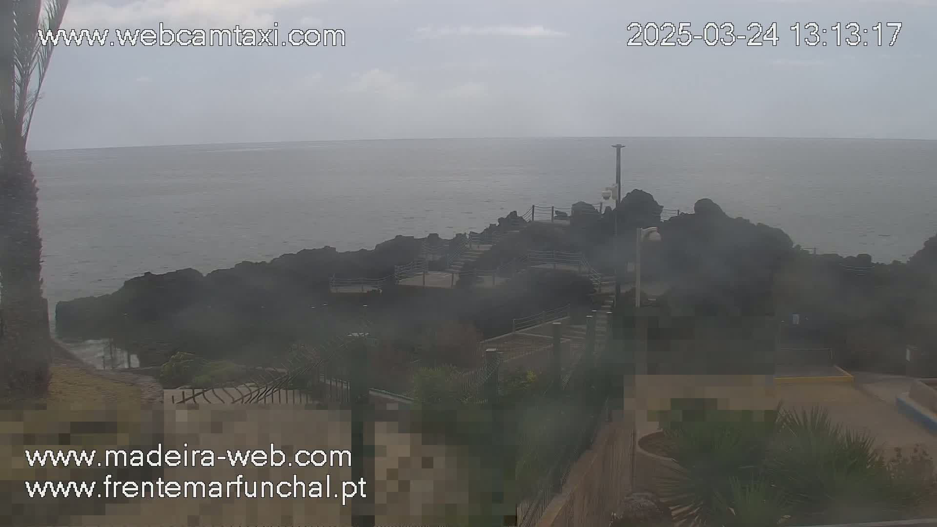 A hazy, overcast day shows a rocky coastline meeting a calm ocean, with some landscaping and structures visible in the foreground.