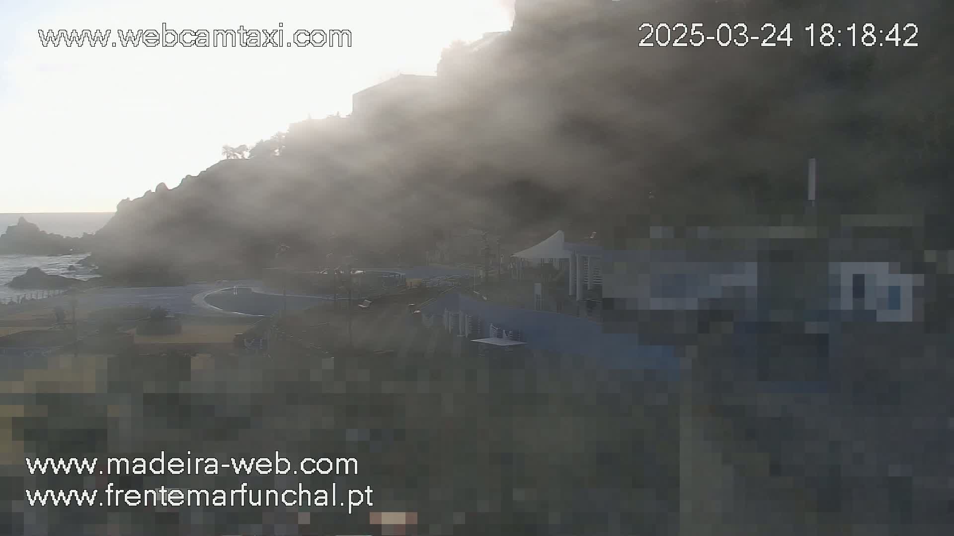 A foggy coastal scene shows buildings and a roadway near the ocean at sunrise.