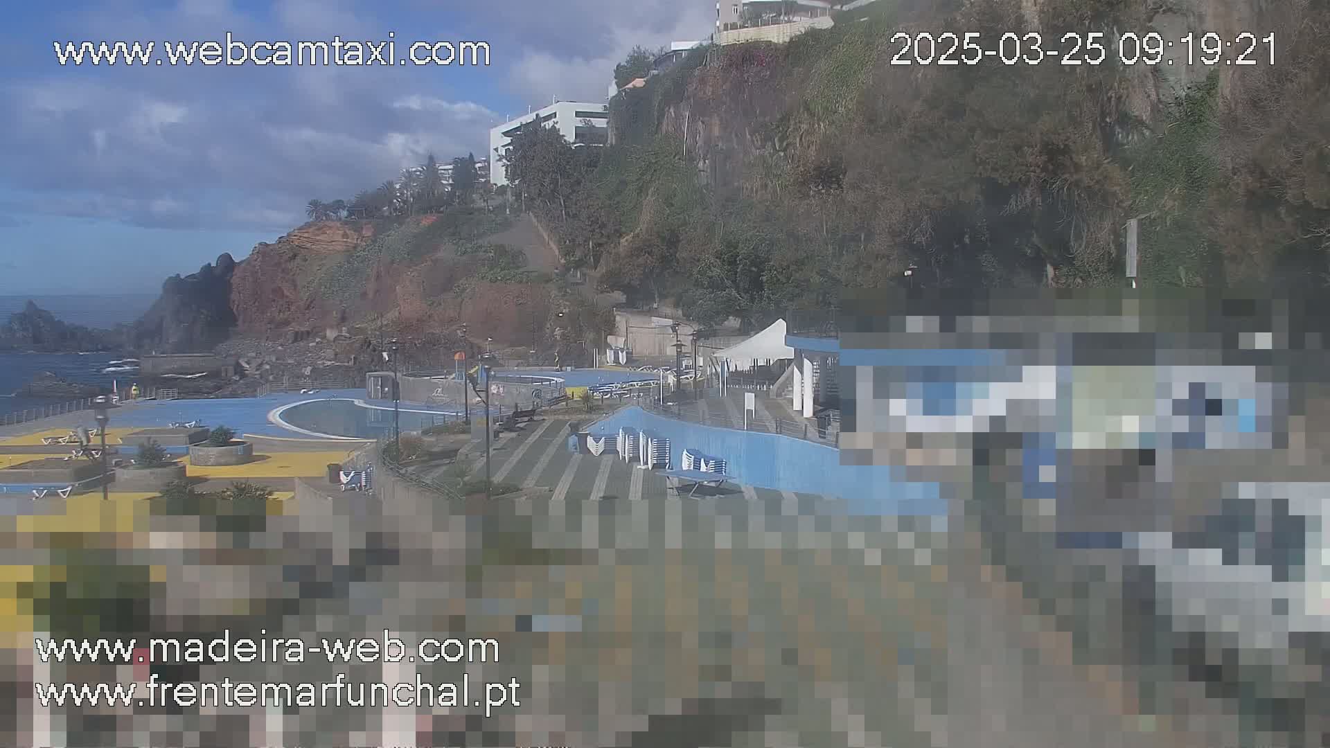 An outdoor view of a seaside pool area on a partly cloudy day, with buildings and cliffs visible in the background.