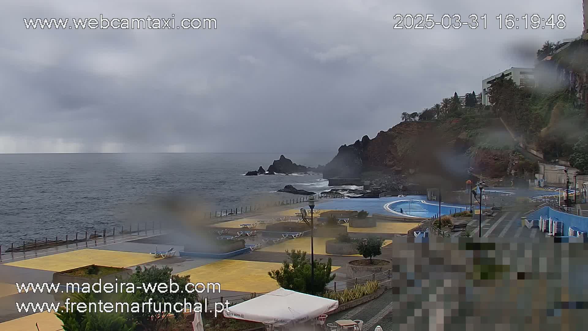 A seaside outdoor pool area is shown on an overcast, rainy day with dark grey skies and a dark blue ocean.