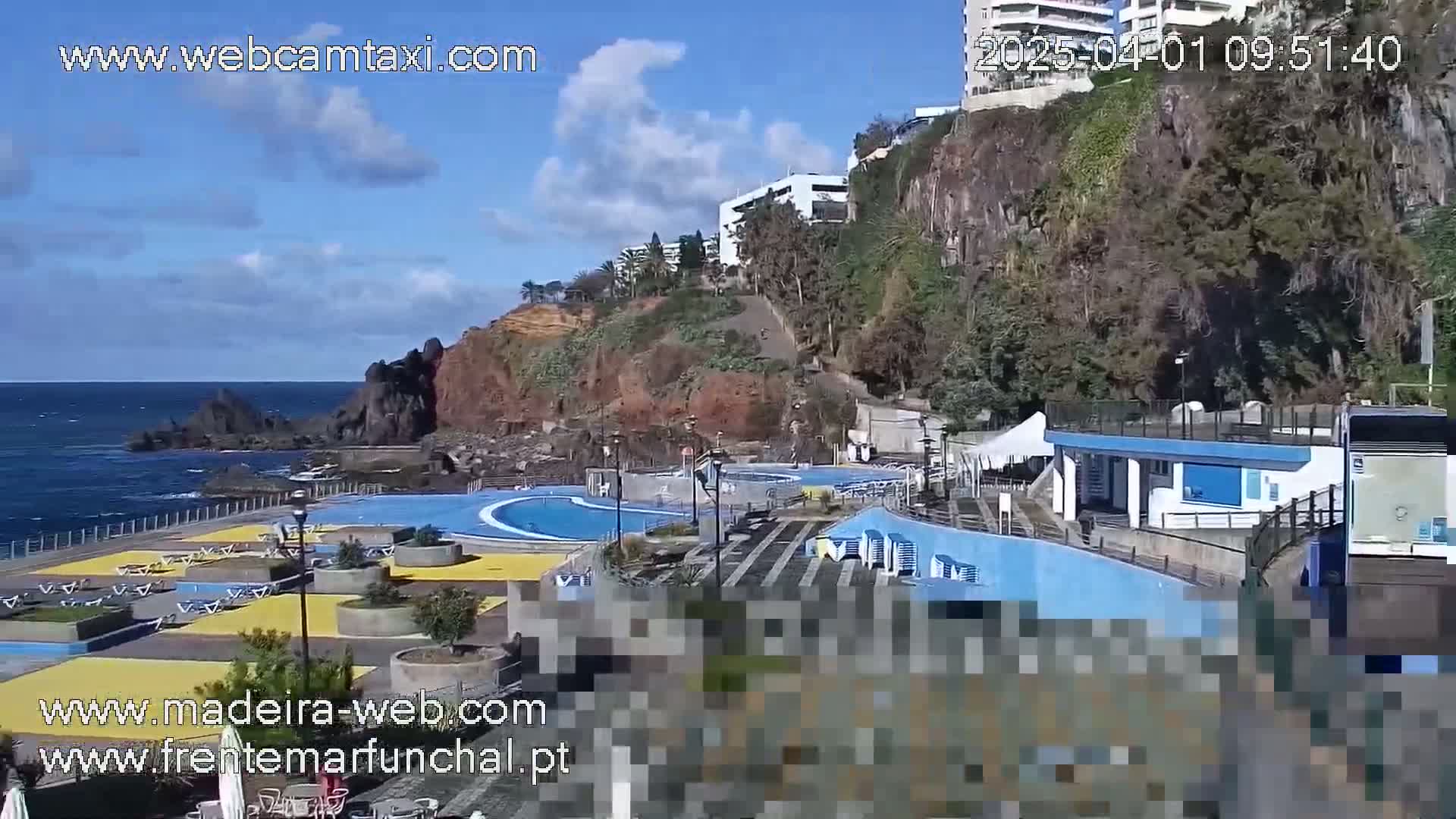 An outdoor oceanside pool complex with a building and stairs sits on a rocky cliff overlooking a calm ocean under a partly cloudy sky.