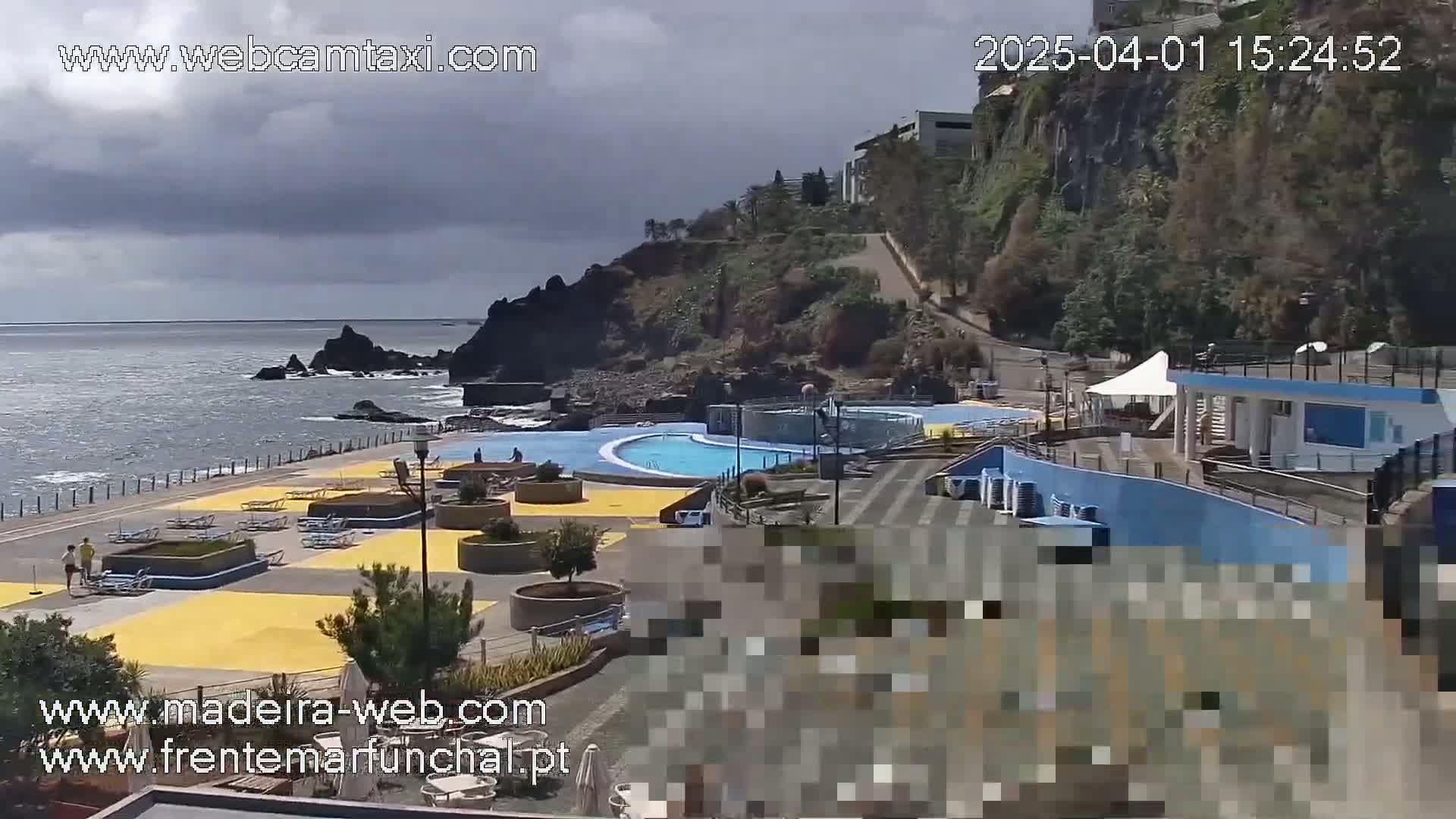 An outdoor oceanside scene shows a swimming pool complex, a paved area with lounge chairs, and a rocky coastline under a partly cloudy sky.
