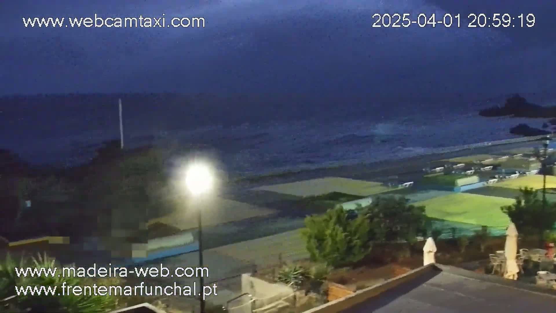 A nighttime view of a dark ocean with waves crashing on a beach near buildings and vegetation under a mostly clear, dark sky.