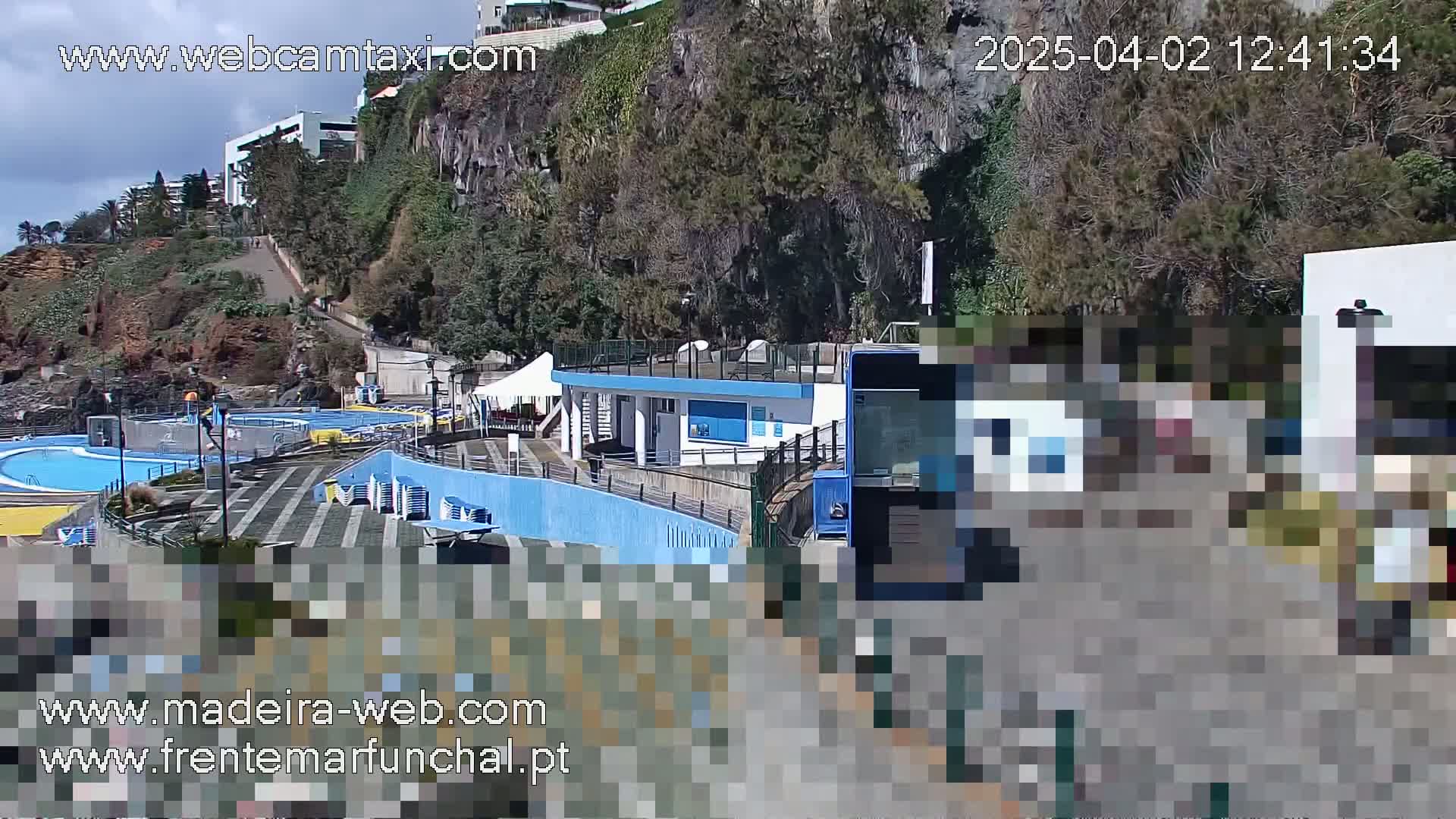An outdoor scene on a partly cloudy day shows a blue swimming pool complex next to a cliffside with buildings and vegetation, and a blue bus partially obscuring the view.