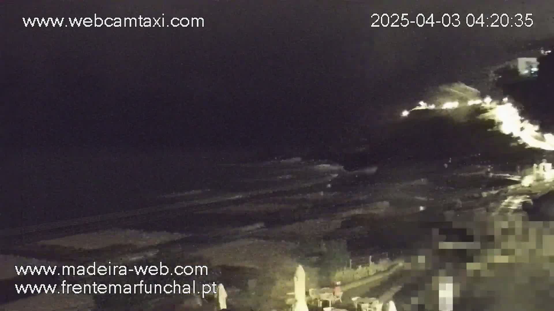 A nighttime coastal scene shows dark ocean waves crashing on a shore, with some lights visible on land and a bright area in the upper right corner possibly indicating light reflecting off the water.